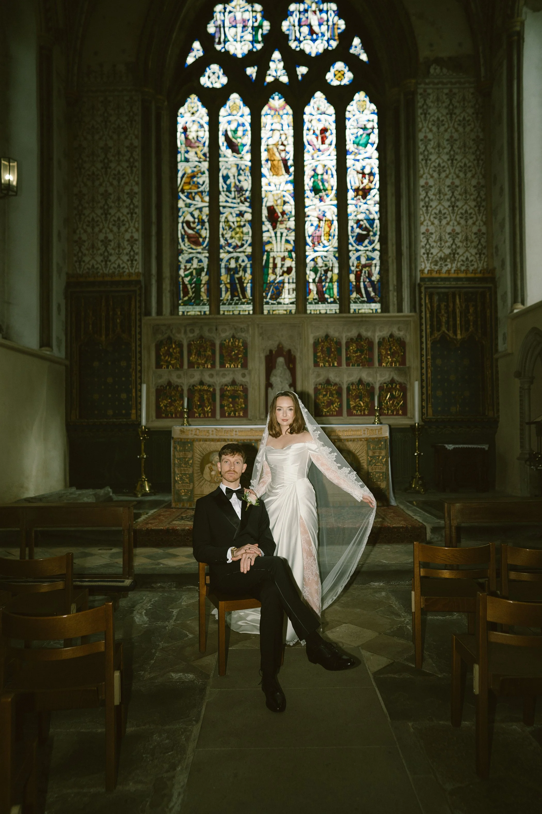 A bride and groom in wedding attire pose inside a church with stained glass windows in the background. The groom is sitting on a chair while the bride is standing behind him, holding his hand and wearing a long veil and white gown.