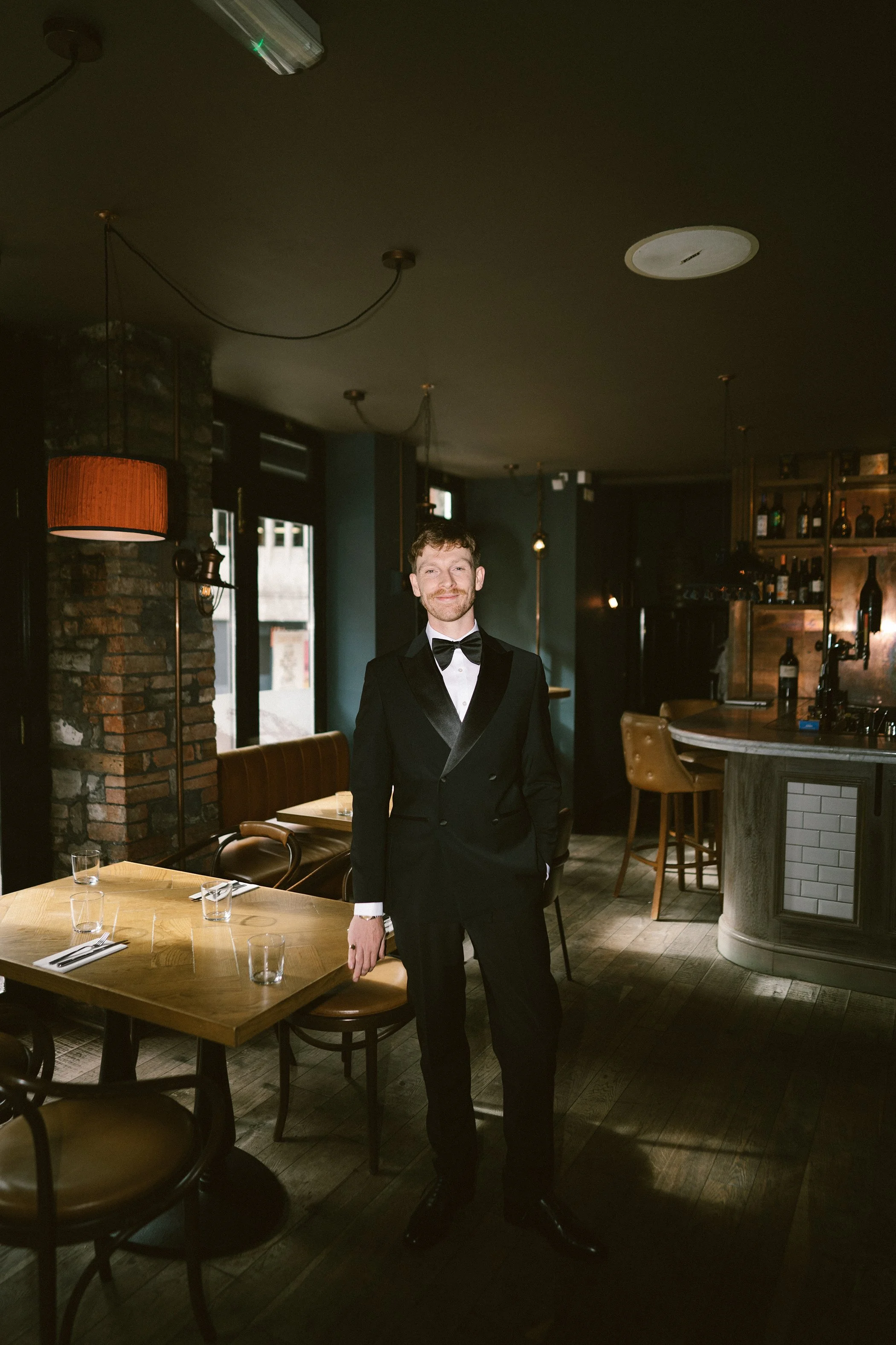 A man dressed in a black tuxedo with a bow tie stands inside a dimly lit restaurant. The restaurant features wooden tables set with glasses and utensils and a counter on the right side with bottles on display. The room has dark walls, some brick acce