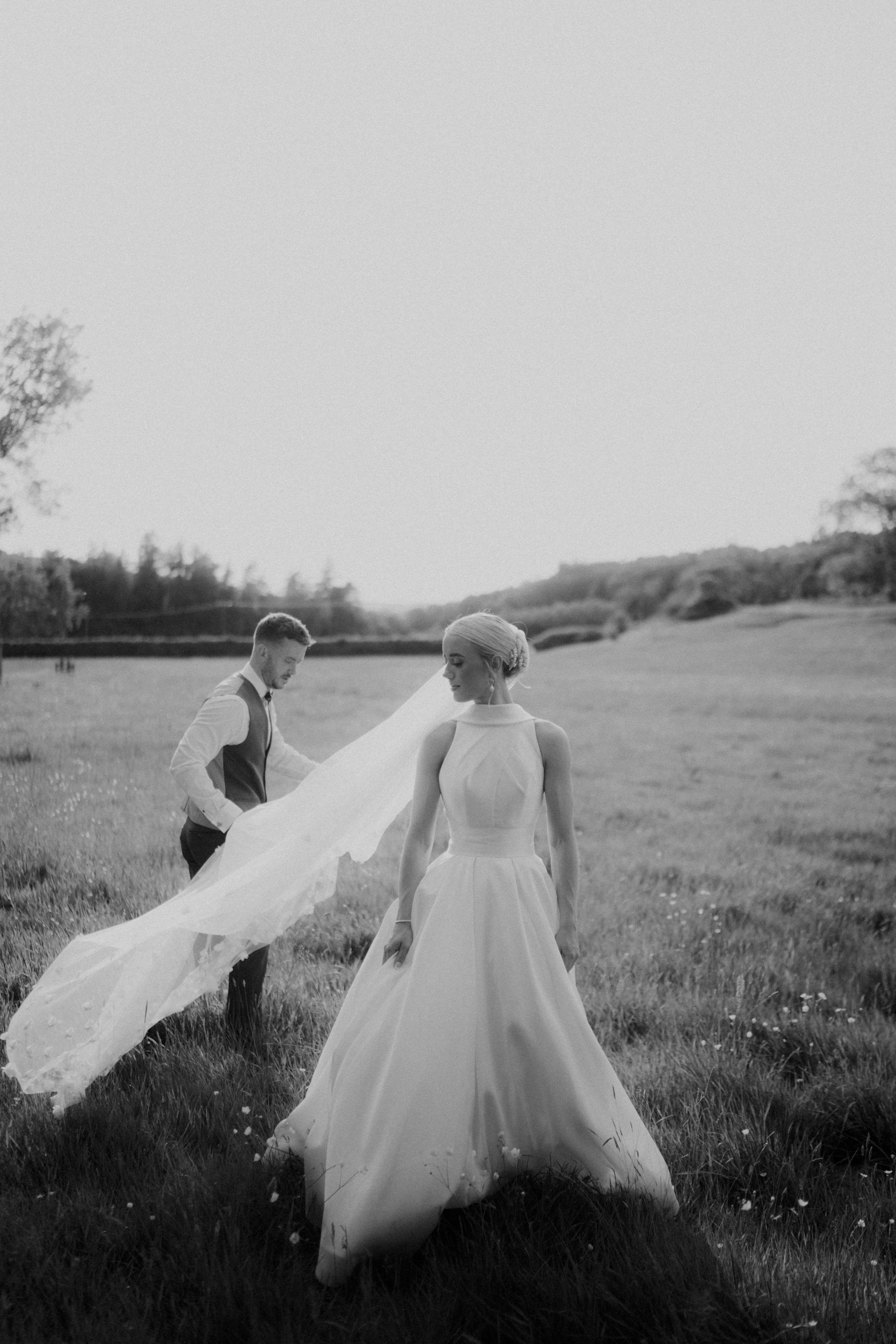 A black-and-white photo of a bride in a long wedding gown with a veil, standing in a field with a groom in a vest and tie behind her, holding the edge of her veil.