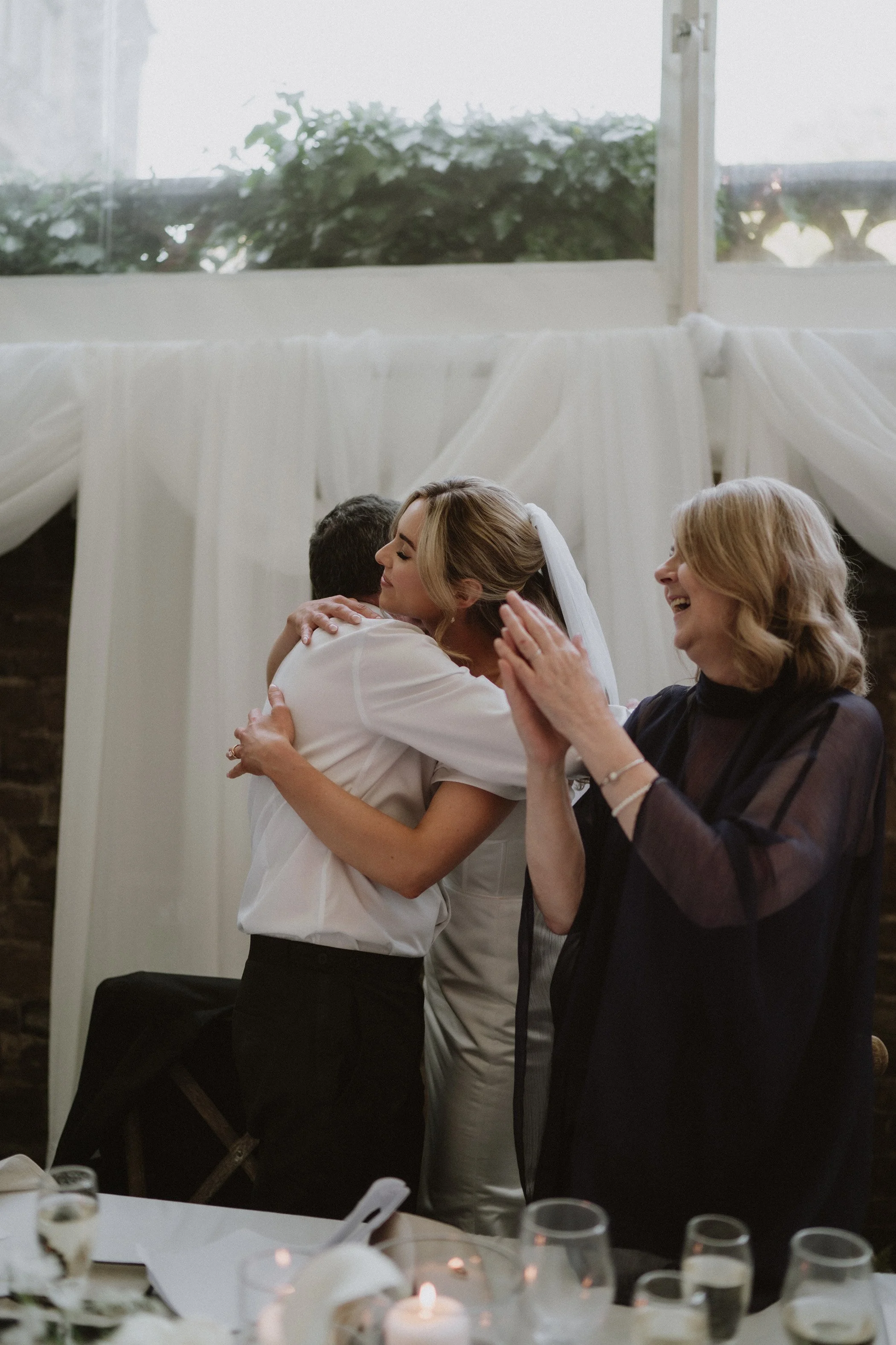 A bride hugging a man at a wedding reception with a woman clapping beside them, all smiling and celebrating indoors.