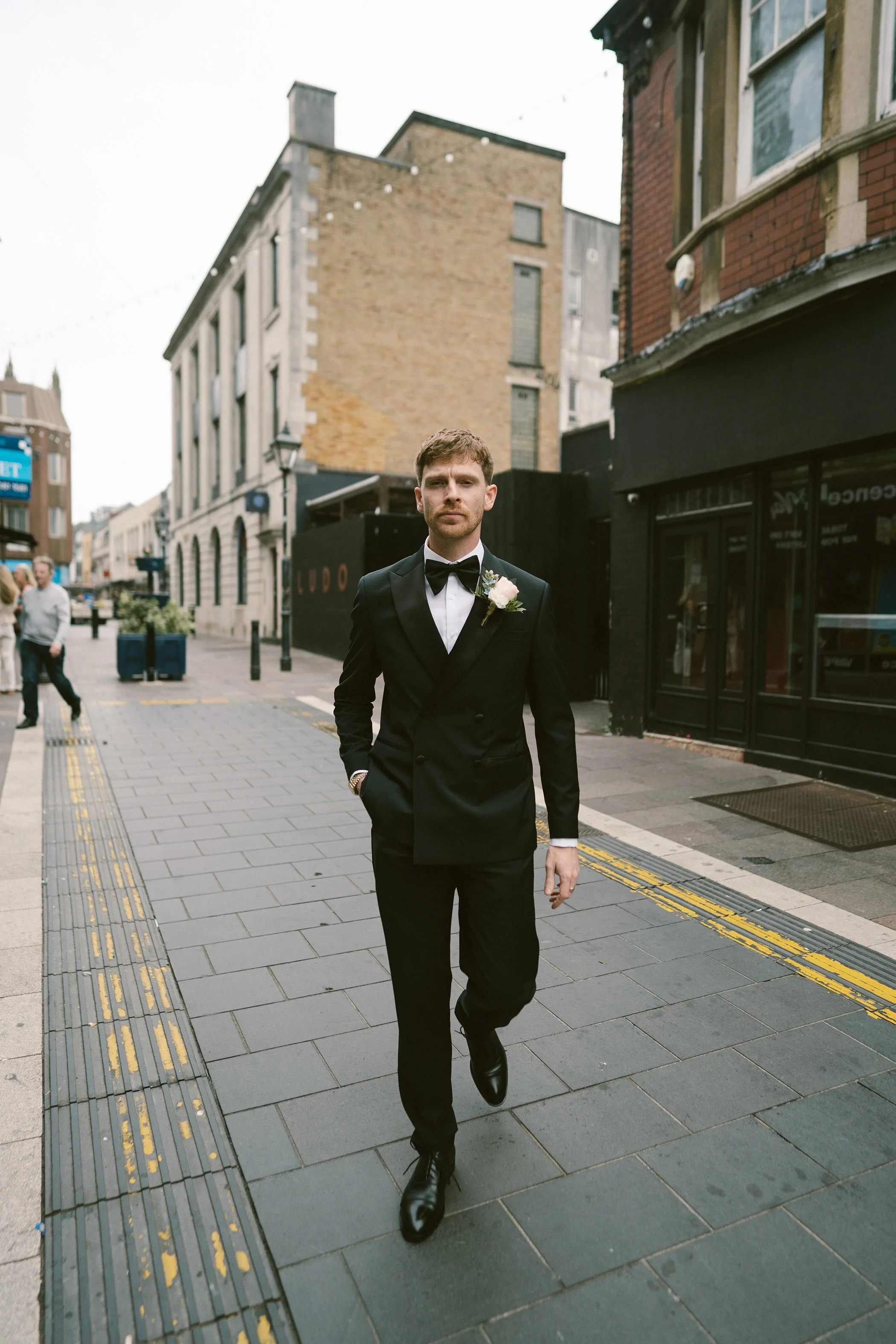 A young man in a black tuxedo with a bow tie and boutonniere is walking confidently down a city street with brick and stone buildings, some with shopfronts, and a few pedestrians in the background.