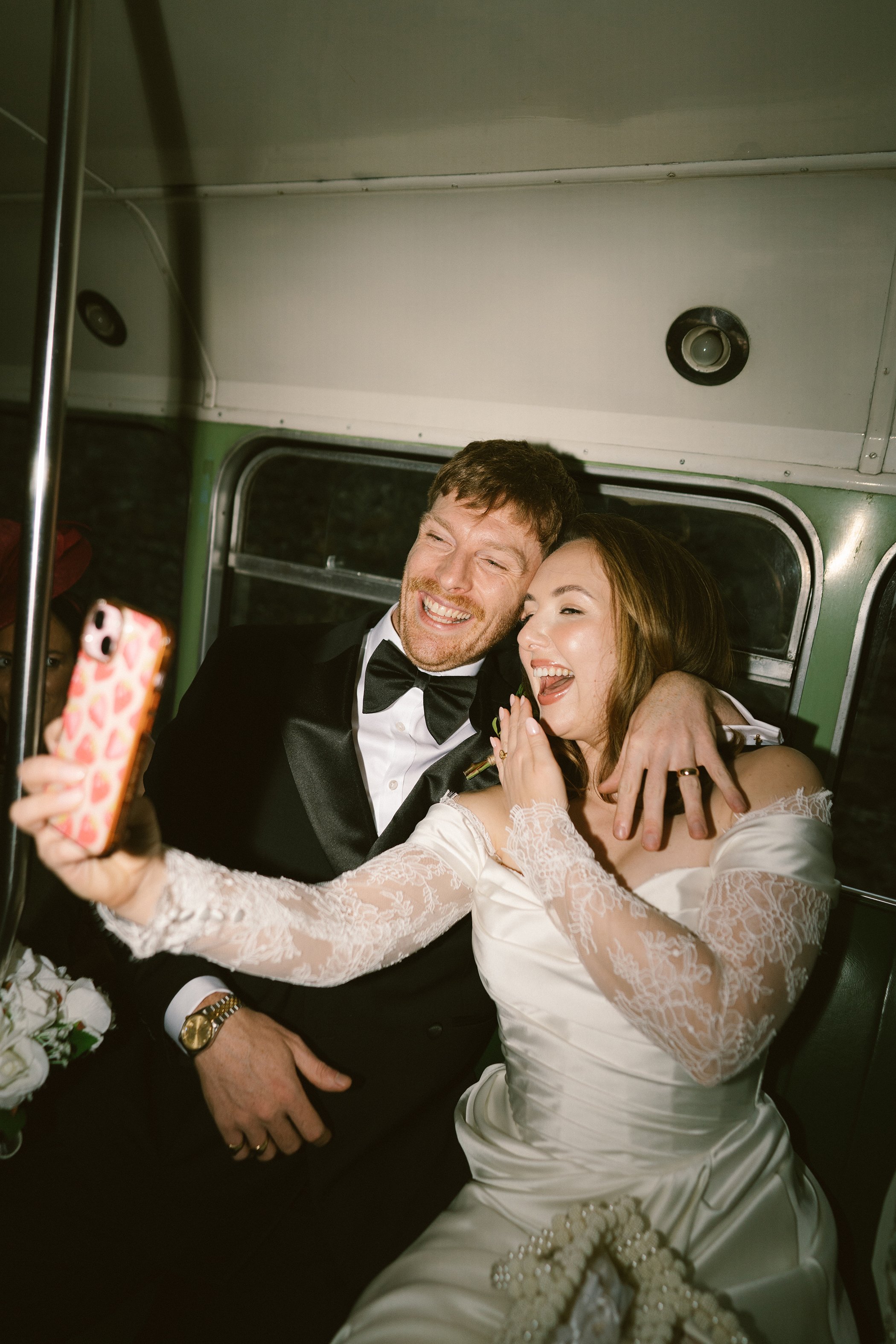 Couple dressed in wedding attire taking a selfie together on a bus, with the bride holding the phone.