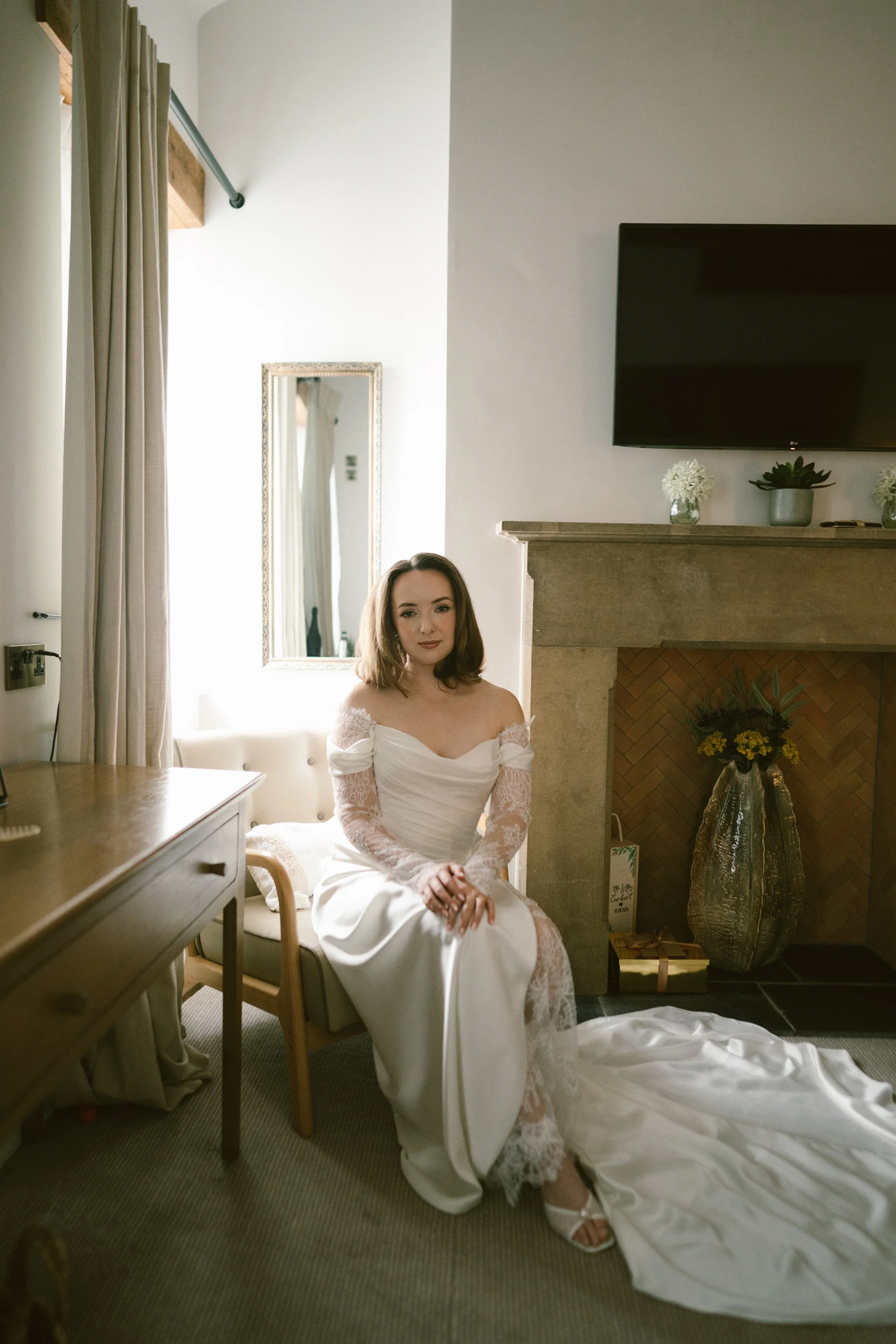 A woman in a white wedding dress sits on a chair in a room with a fireplace, a large television, and decorative flowers.