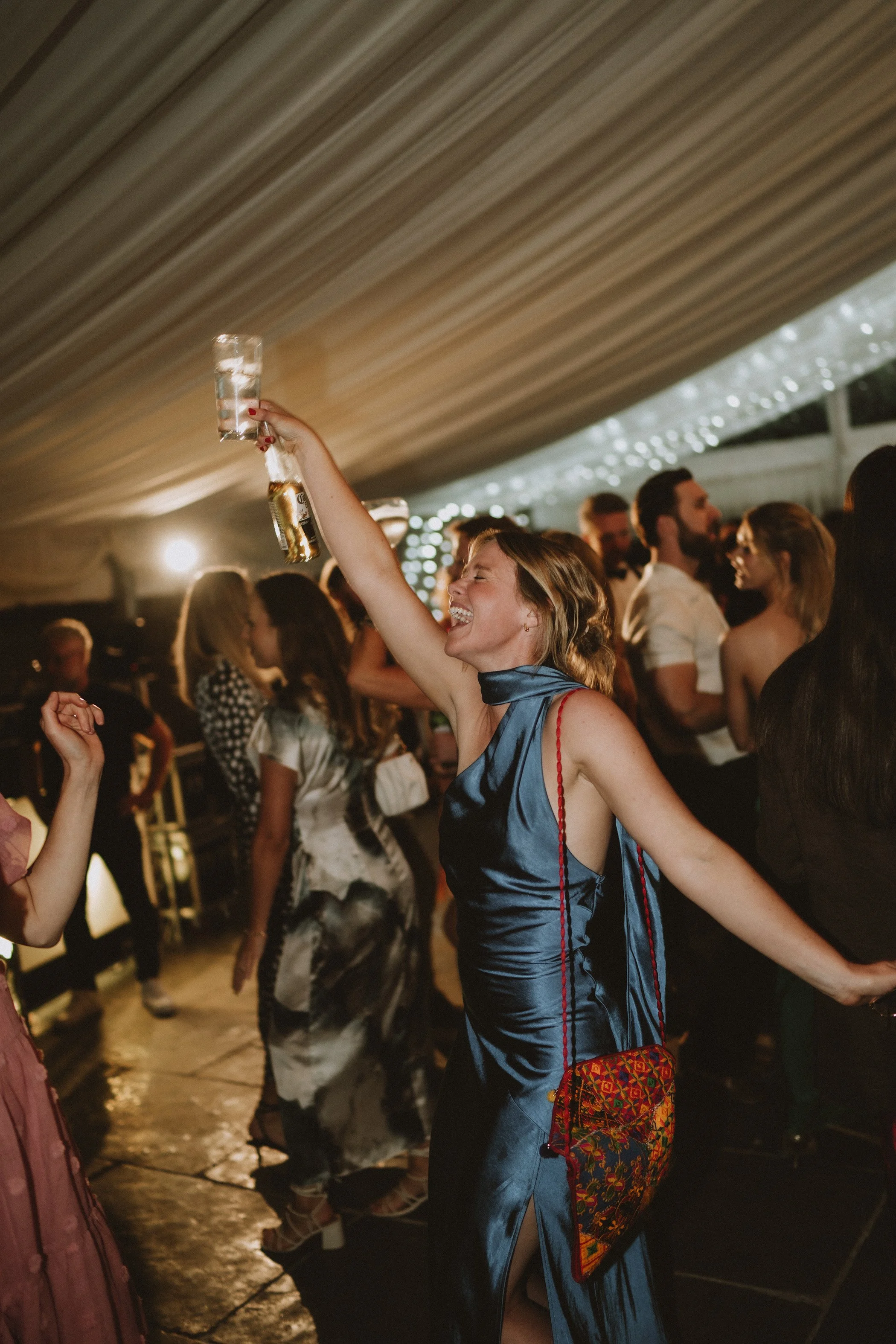 A woman in a blue satin dress cheers with a glass in hand at a lively indoor party, surrounded by dancing people.