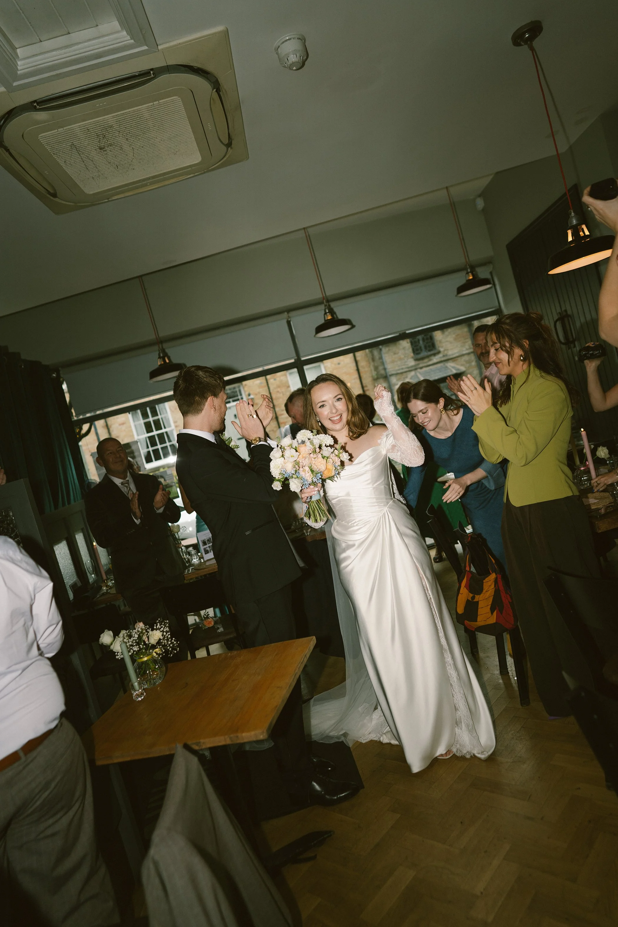 A bride in a white wedding dress holding a bouquet of flowers, surrounded by smiling friends and well-wishers indoors during a wedding celebration.