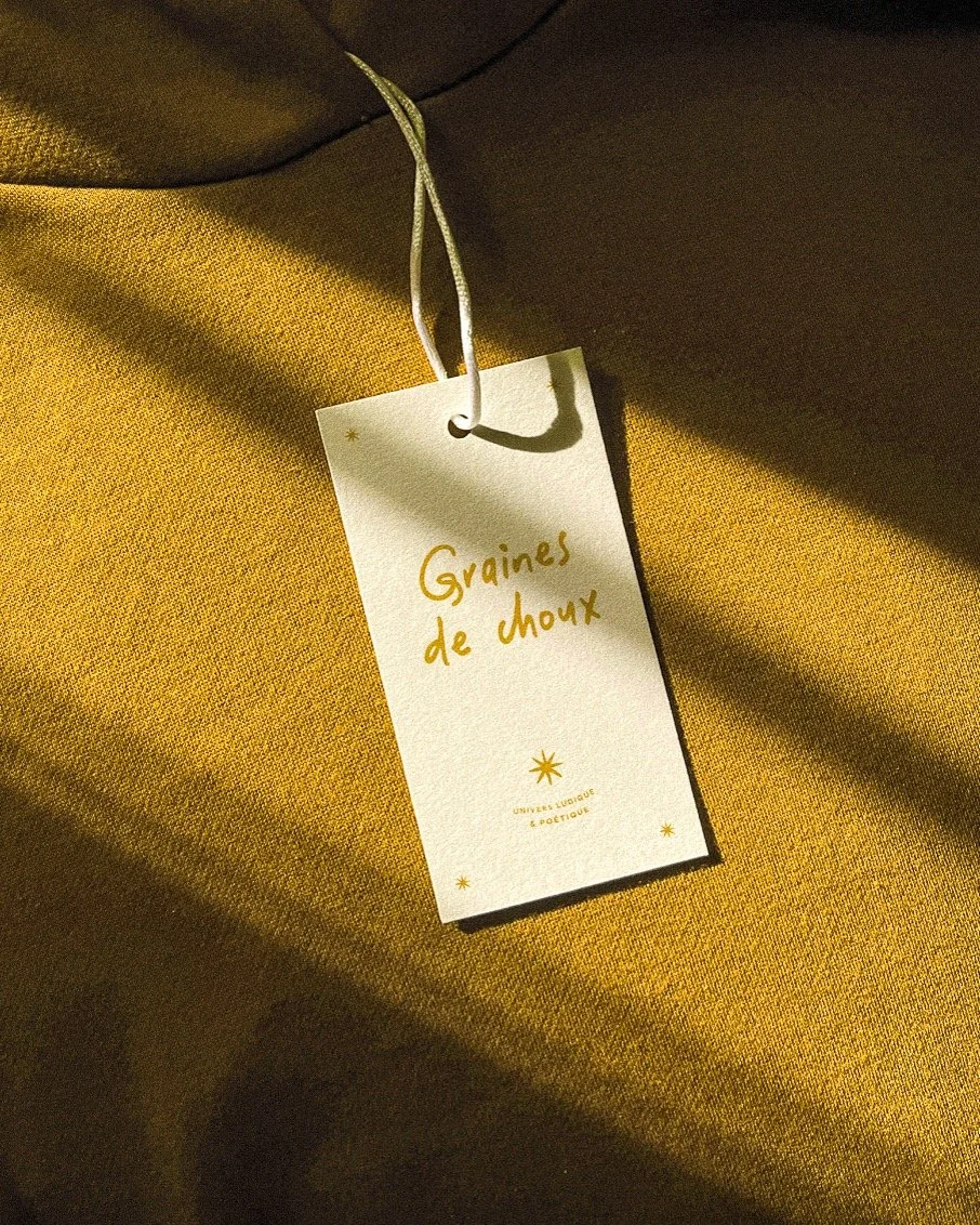 Étiquette blanche avec le texte "Graines de choux" écrit en orange, attachée à un tissu jaune avec une cordelette blanche.