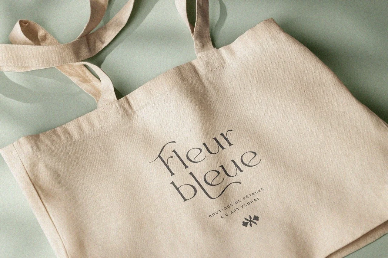 Sac en toile beige avec l'inscription 'fleur bleue' et une sous-phrase 'boutique de pétales & d'art floral' en lettres noires, posé sur un fond bleu clair.