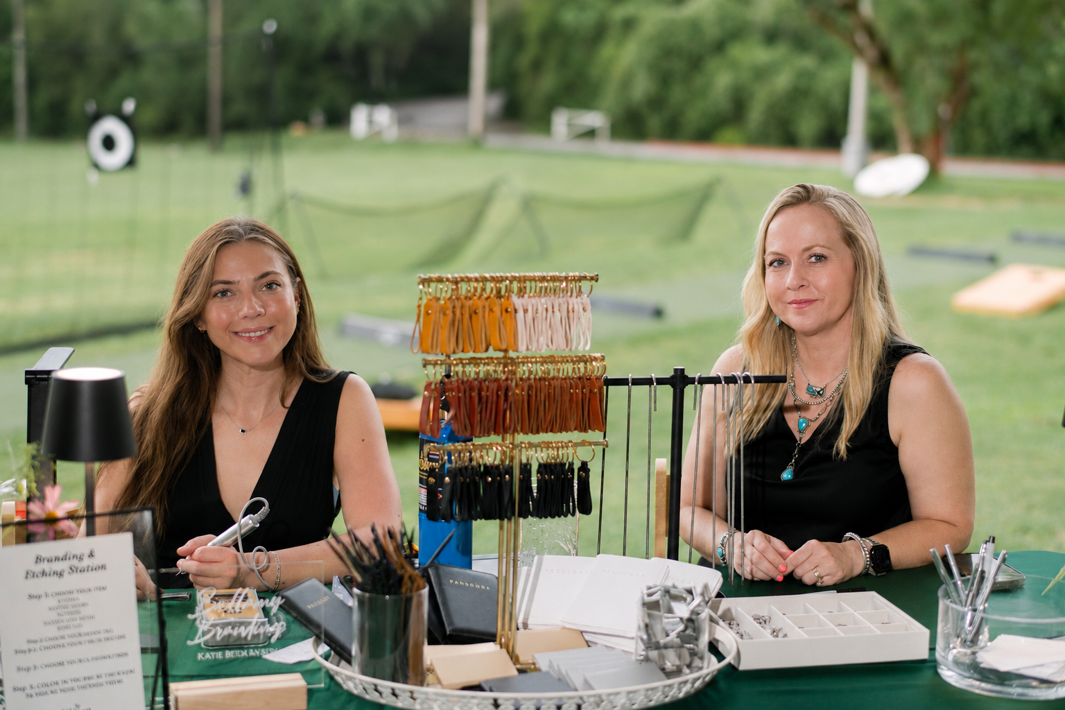 Two women sitting at a jewelry stand outdoors with trees in the background, displaying jewelry and accessories.