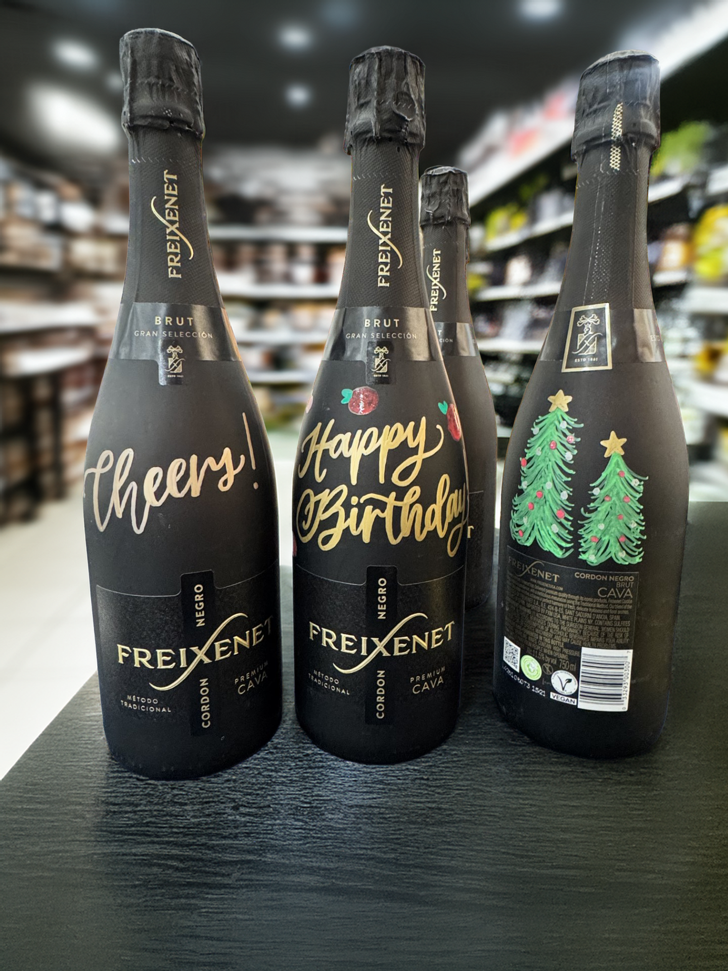 Three bottles of Freixenet sparkling wine on a black surface in a grocery store, decorated for celebration with one bottle saying 'Cheers!', another saying 'Happy Birthday,' and the third featuring Christmas trees with ornaments.