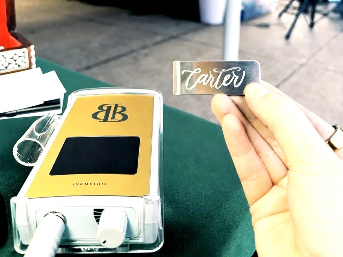 A hand holding a metal sign with the word 'Carter' written in cursive, next to a yellow device with a BB logo and a blank black screen on a green table.