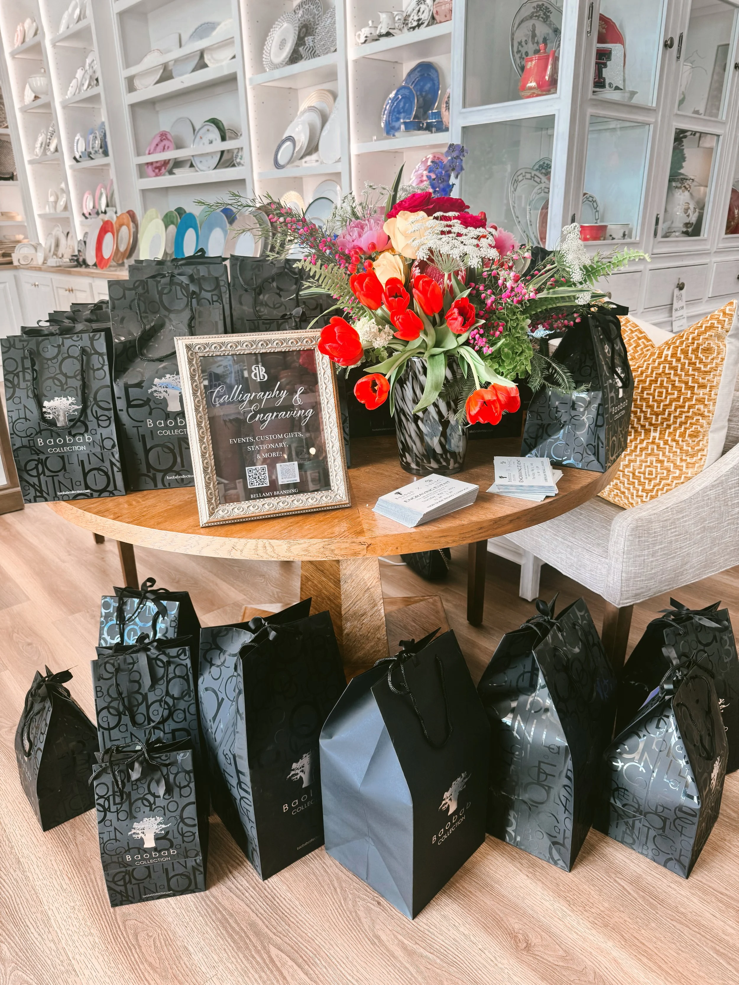 A round wooden table displaying a floral arrangement with red tulips, pink roses, white flowers, and greenery. Around the table, multiple black and blue shopping bags with handles and bows, branded 'Baobab Collection,' are arranged. In the background