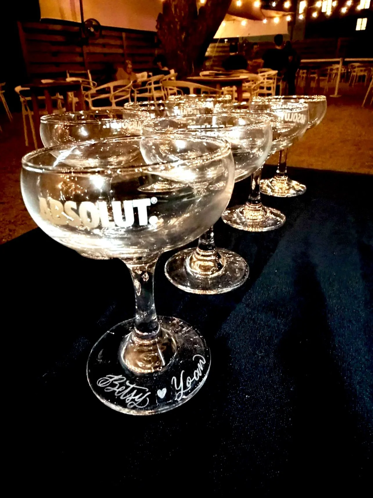 A row of six empty cocktail glasses with the word 'BLOUT.' printed on one, and 'Happy Birthday' written on the base of another, set on a black tablecloth in a dimly lit outdoor venue with wooden chairs and string lights in the background.