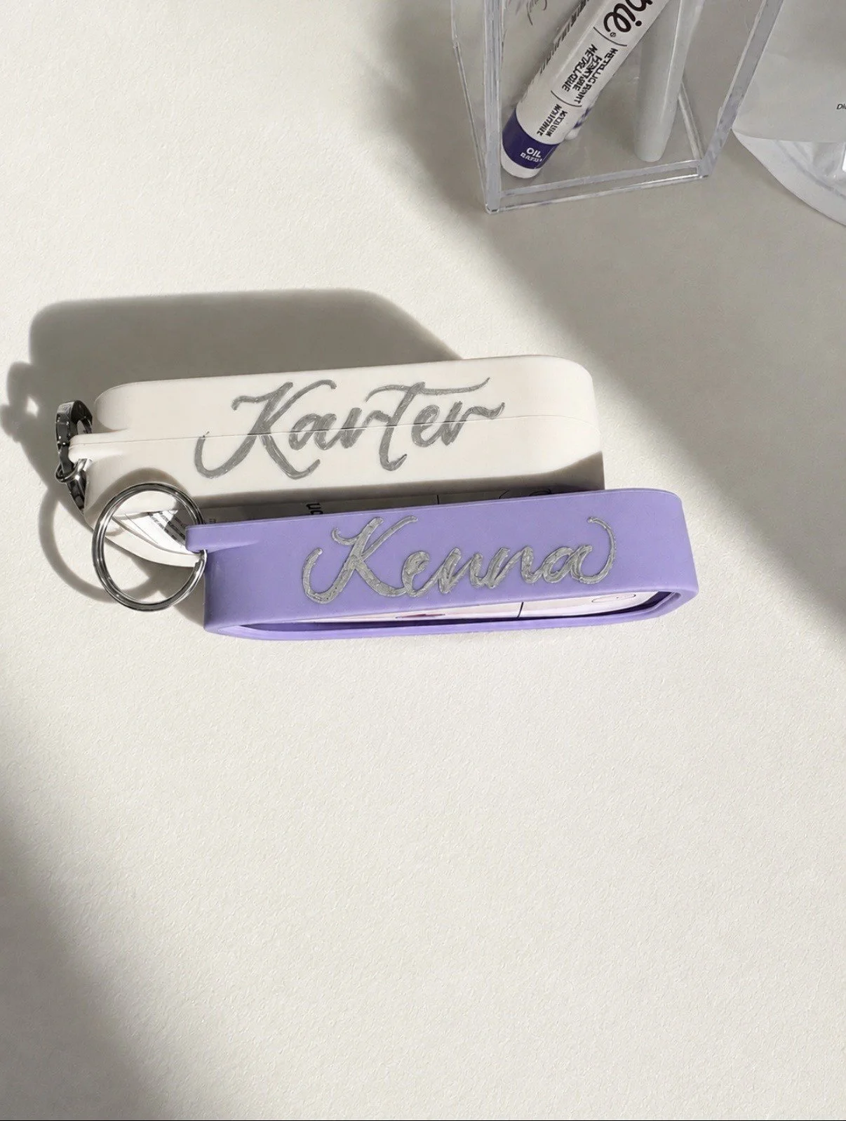 Two keychains, one white and one purple, with the names 'Karter' and 'Kenna' embroidered on them, resting on a white surface. In the background, a clear plastic container holds a white marker and a purple-labeled item.