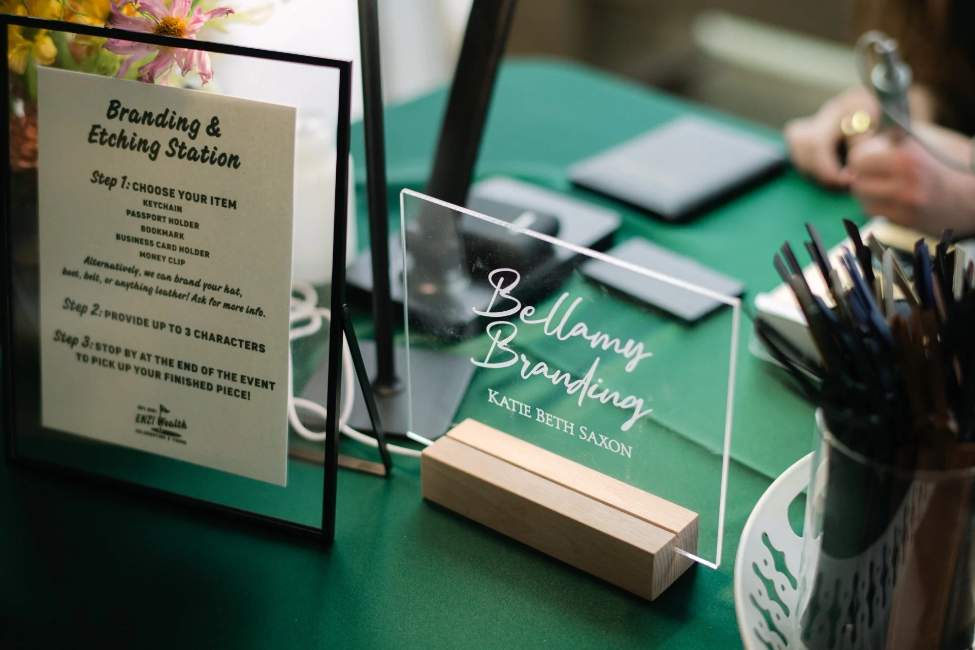 A display table at a branding and etching event with a sign explaining the steps to create personalized items, including a clear acrylic sign with white text that reads 'Bellamy Branding,' a black pen holder with pens, and a small wooden block supporting the sign.