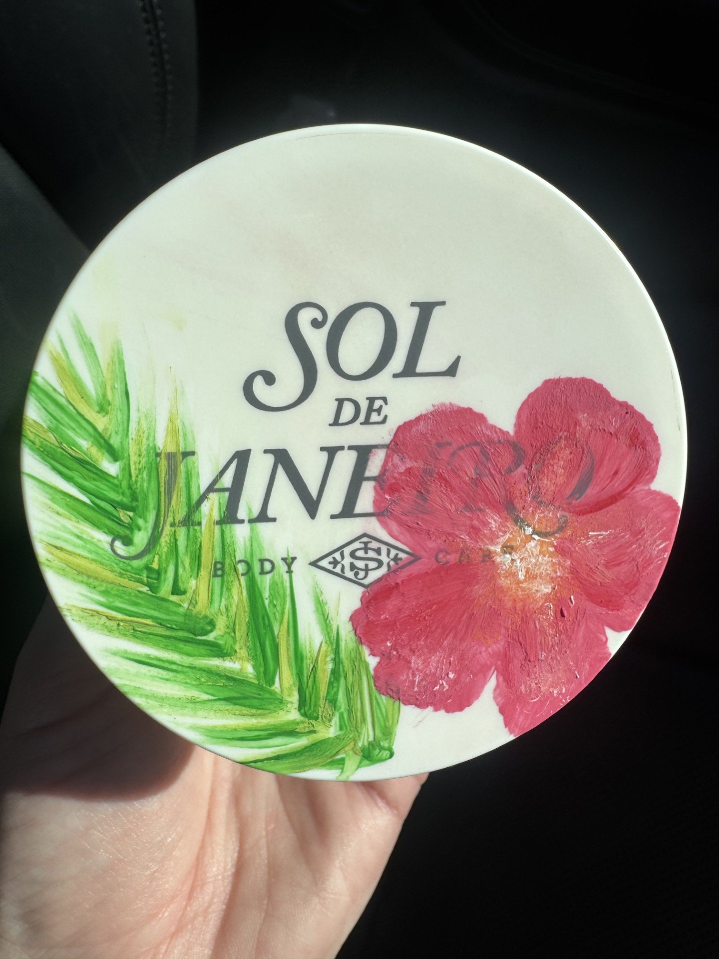 A hand holding a round container with a floral design, featuring a red flower and green leaves, and partially visible text that reads 'SOL DE JANEIRO' and 'BODY'.