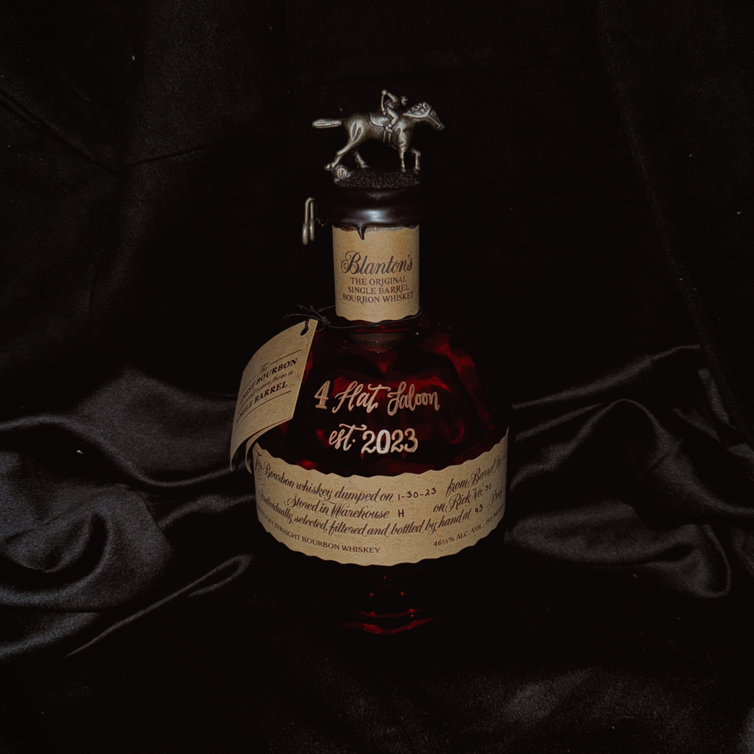 A bottle of Blanton's bourbon whiskey with a metal horseshoe and jockey figurine on top, placed on black satin fabric.