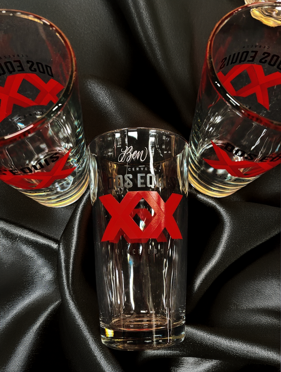 Three glasses with Dos Equis branding on a black fabric background, with the middle glass empty and the two outer glasses partially filled with a clear beverage.