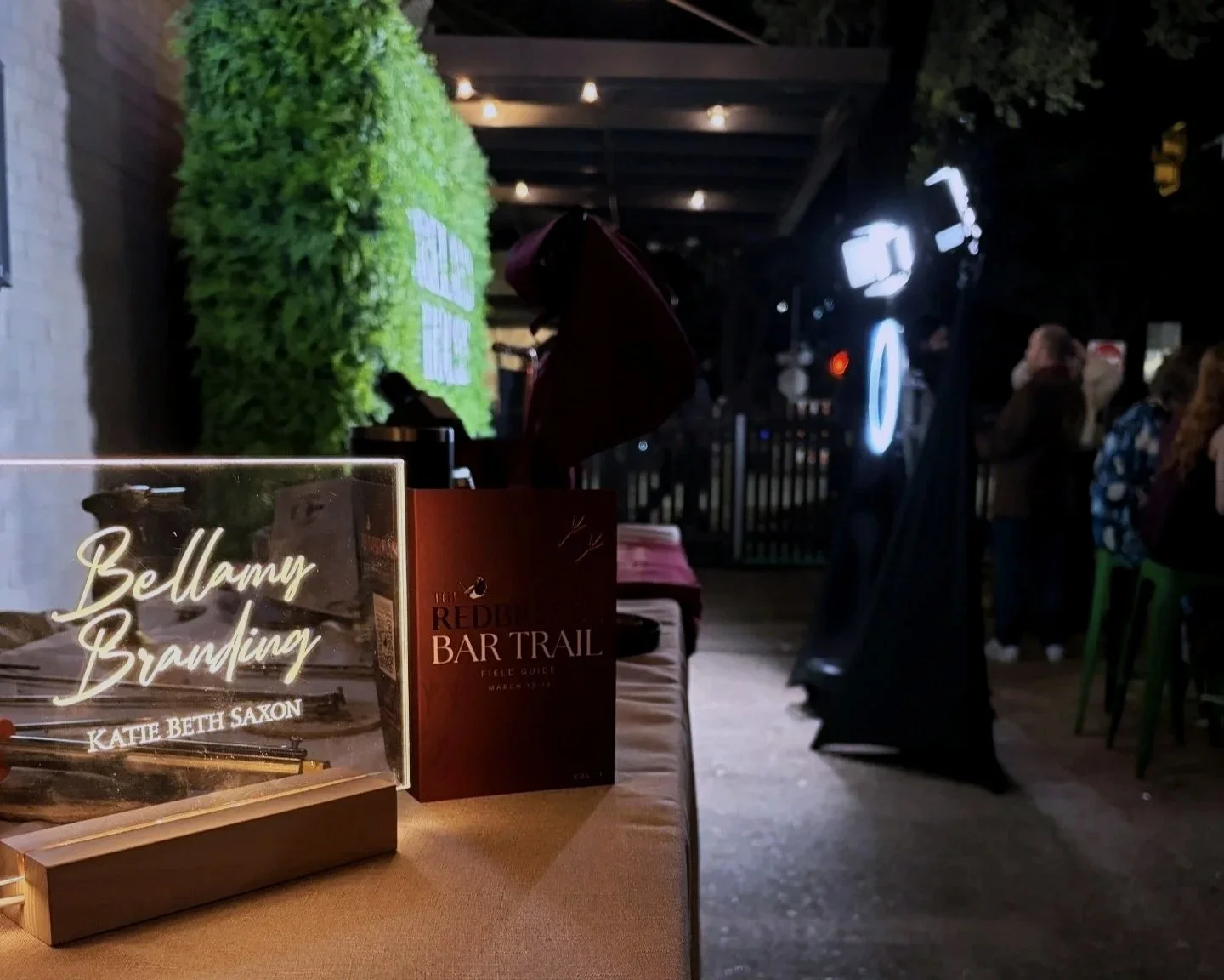 Display table with books, one titled 'Bellamy Branding' by Katie Beth Saxon, and another called 'The Redbird Bar Trail.' In the background, people are standing outside at night near a lit-up sign. There is a line of greenery along the left side and a fence separating the outdoor area.