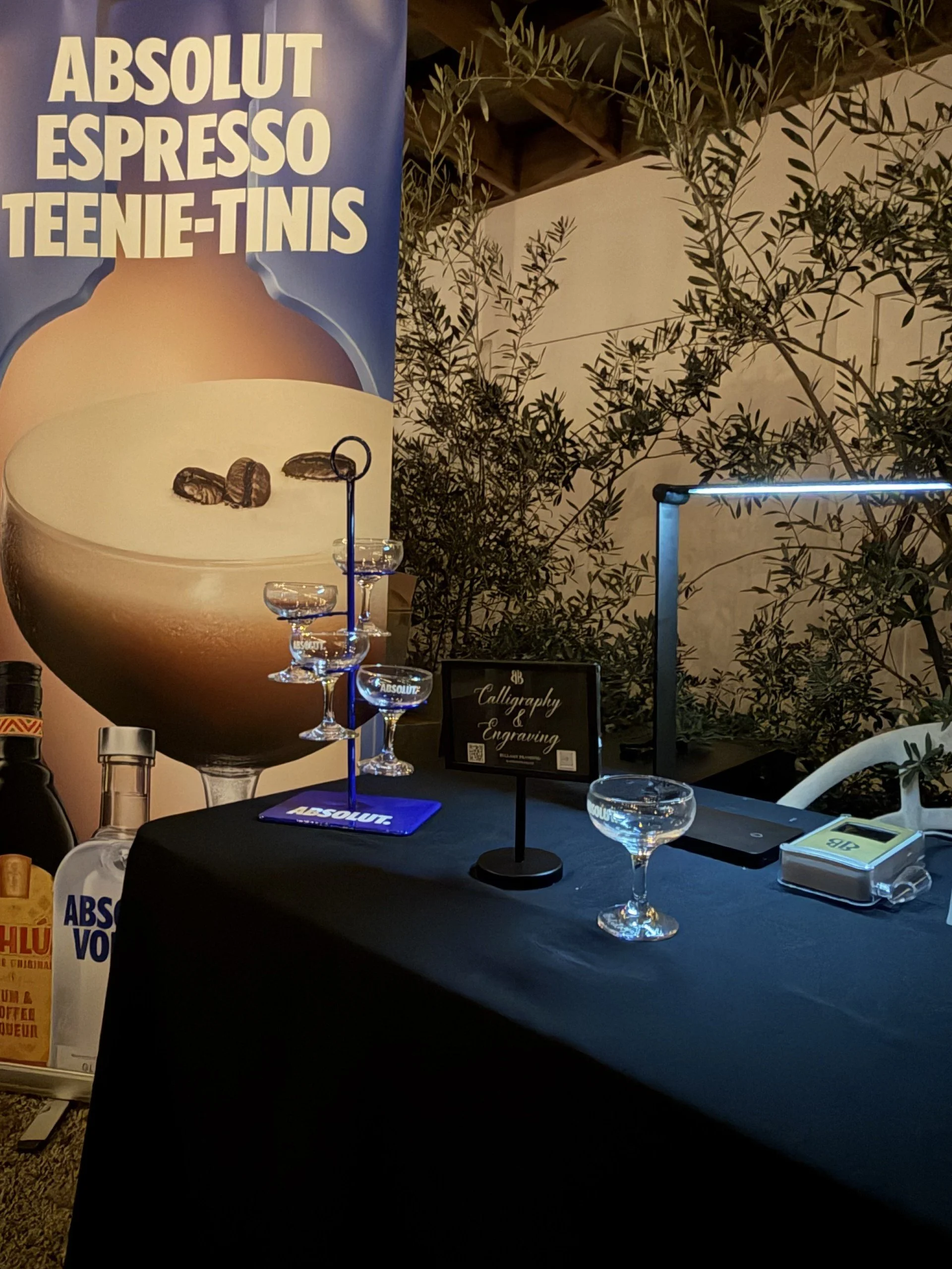 A table with a black tablecloth displaying empty glassware, a small blackstand with the text "Calligraphy & Engraving," and promotional signs for ABSOLUT vodka. Behind the table, there is a large banner with an iced coffee in a glass, coffee beans, a