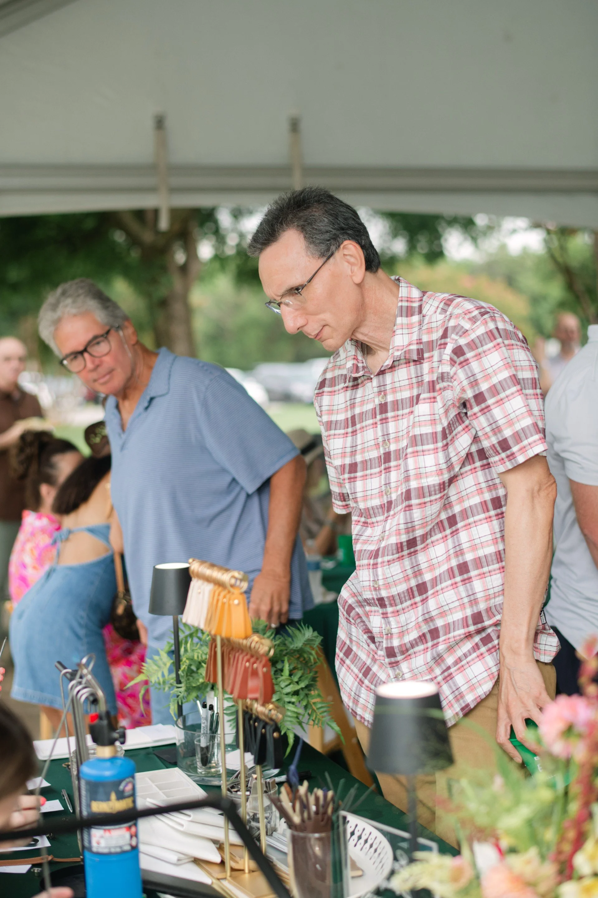 Two men at a market stall, examining items, surrounded by plants and various objects, outdoors with trees in the background.