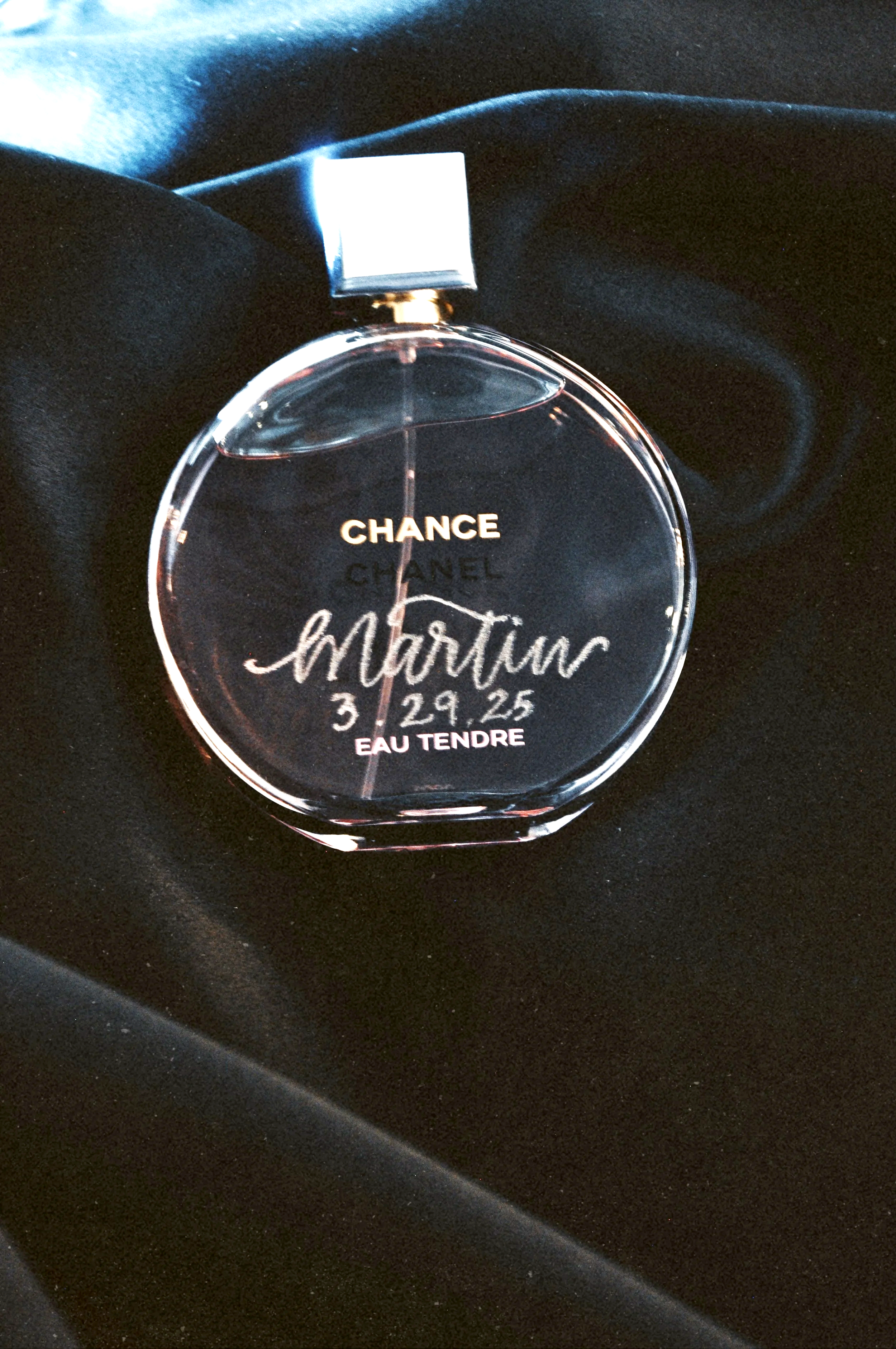 A clear Chanel Chance perfume bottle with personalized engravings, resting on black fabric.