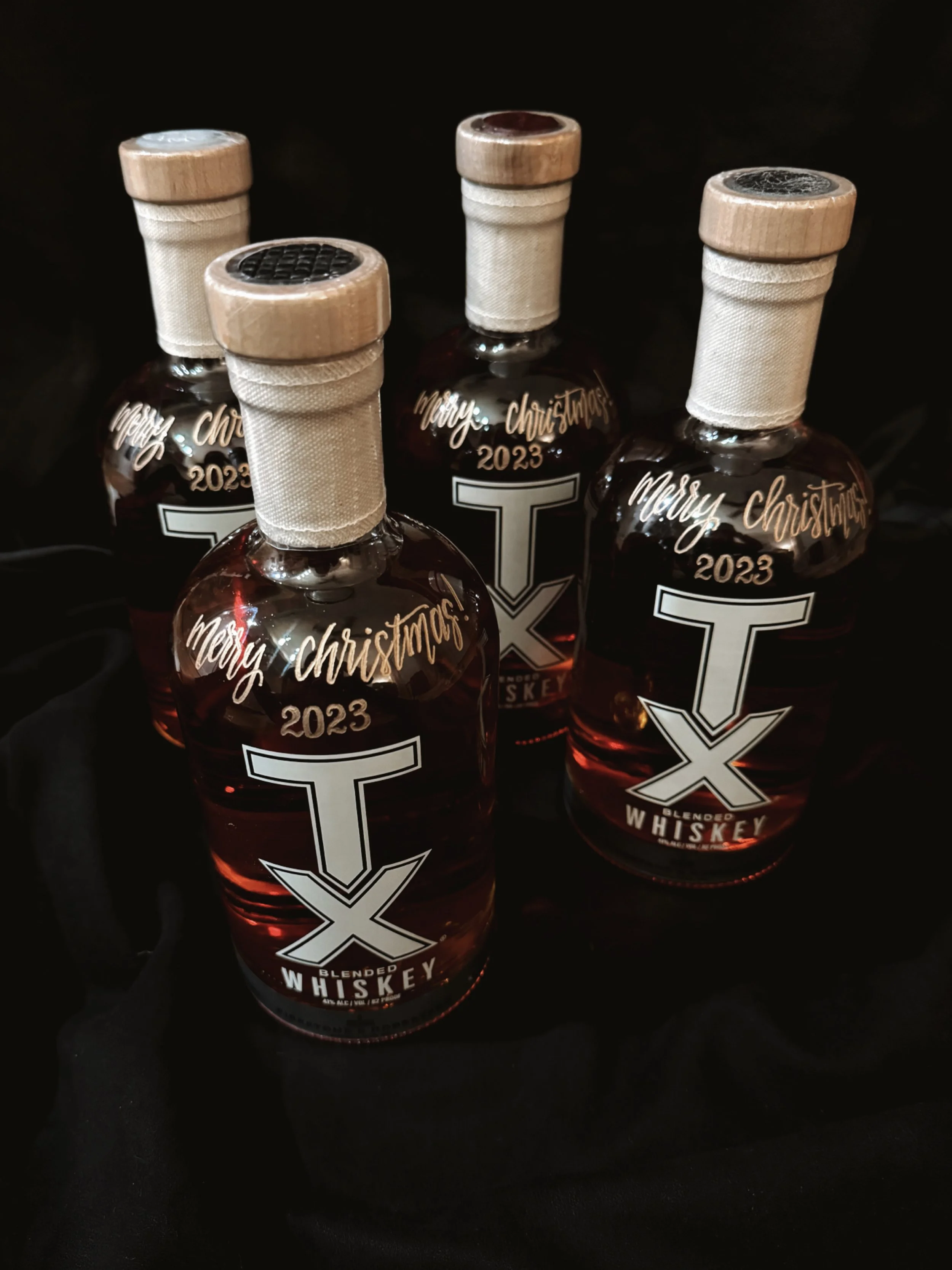 Four bottles of blended whiskey with a Christmas label that reads "Merry Christmas 2023" and a large white T logo on the front, set against a black background.