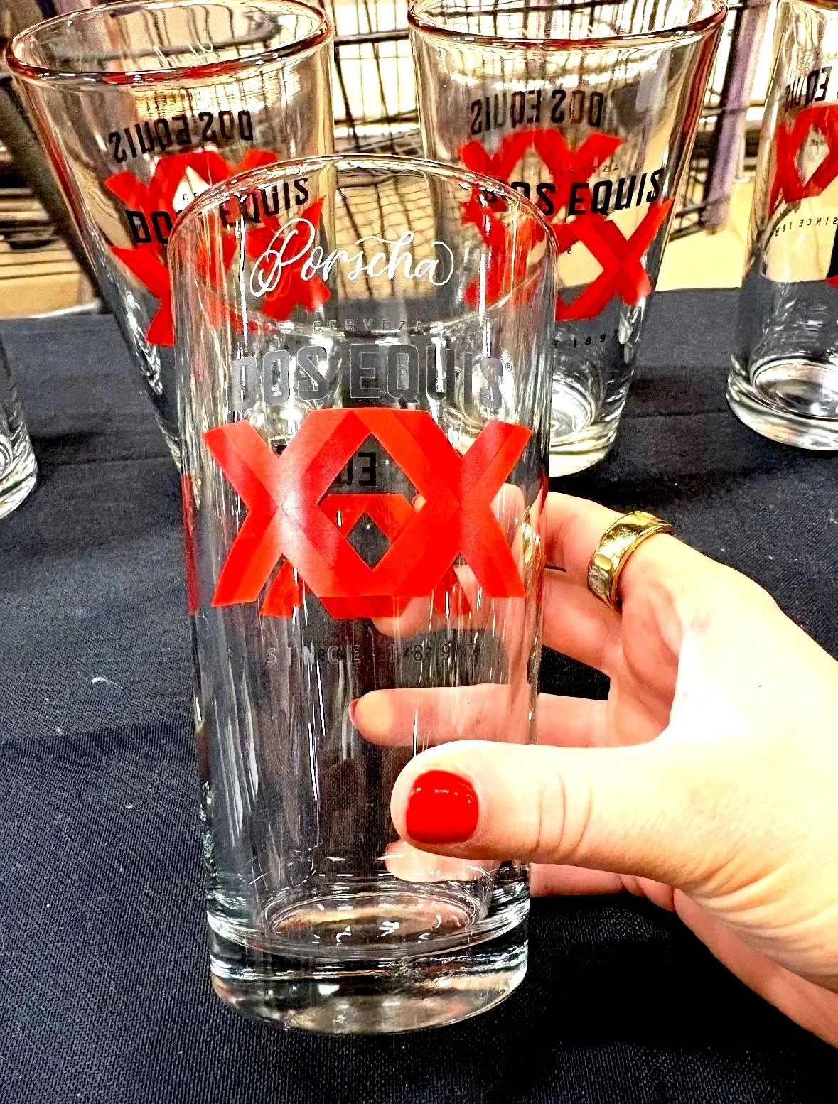 Clear pint glass with red and white branding that reads 'Porcha' and 'Dos Equis' with red 'X' logos, held by a hand with red painted nails and a gold ring, with other similar glasses in the background on a black surface.