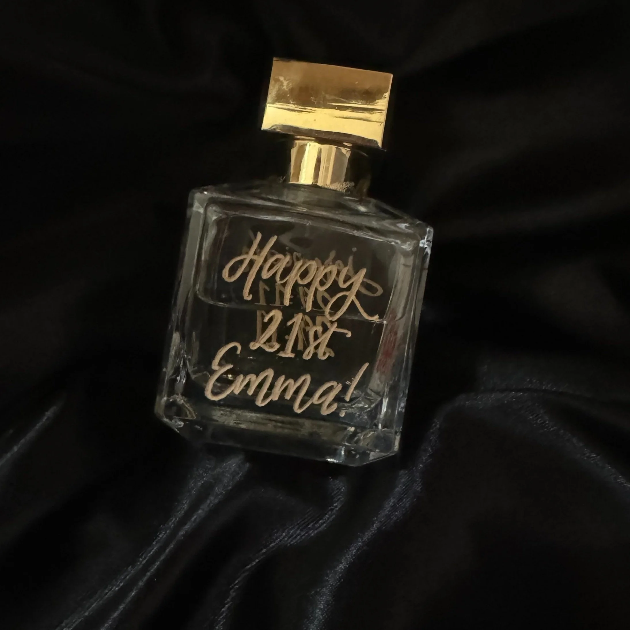 A glass perfume bottle with a gold cap, engraved with a birthday message for Emma on black fabric background.