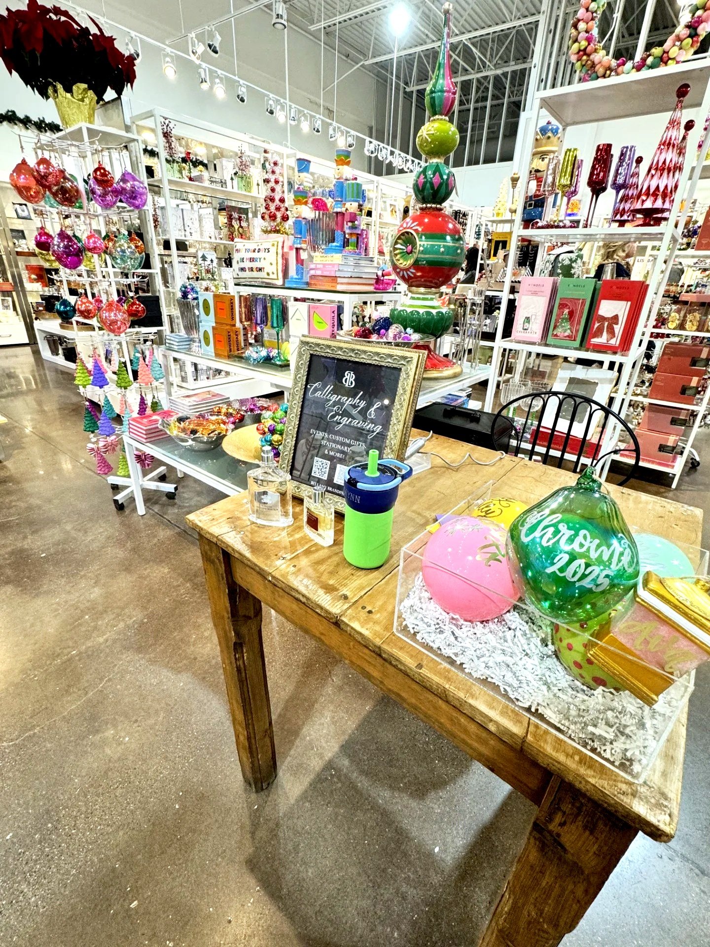Display of colorful holiday ornaments and decorations in a store, including ornaments, books, and a chalkboard sign, with a wooden table holding balloons and a drink container.