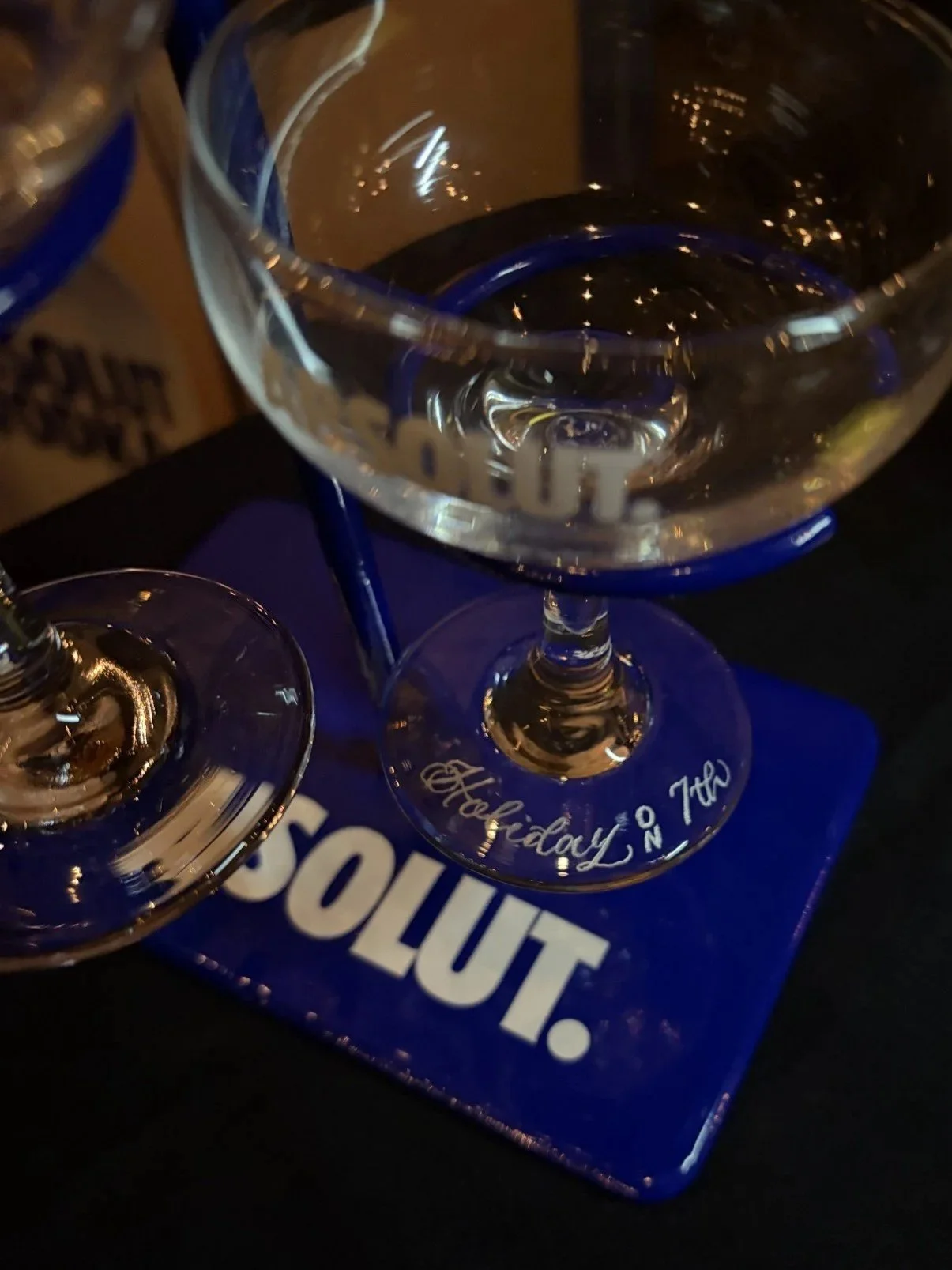 Two empty cocktail glasses on a blue tray with the words "Happy Hour" written on it. One glass has the word "Holiday" written on the stem. The tray is on a dark surface.