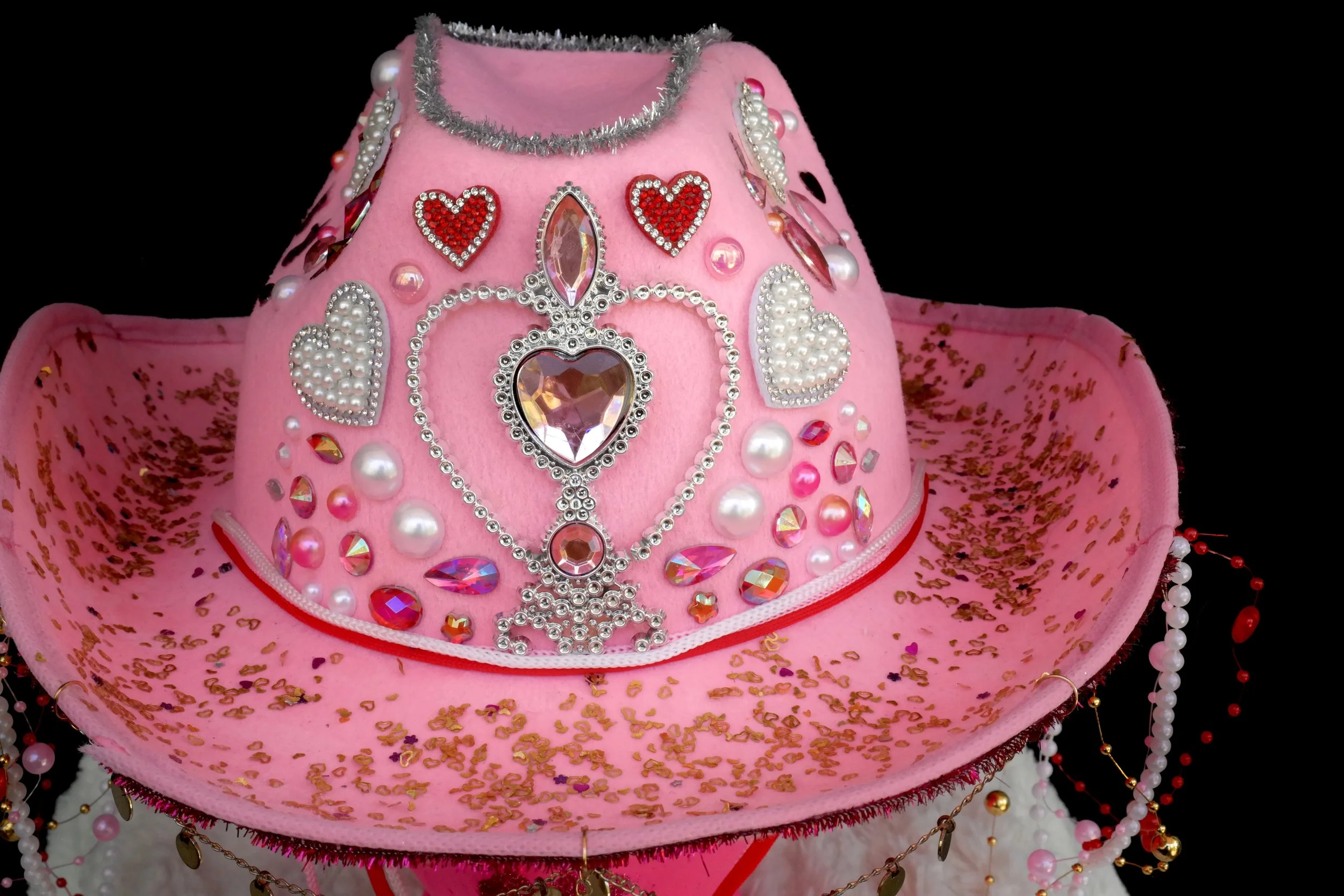 Pink cowboy hat decorated with rhinestones, pearls, hearts, and jewels, set against a black background.