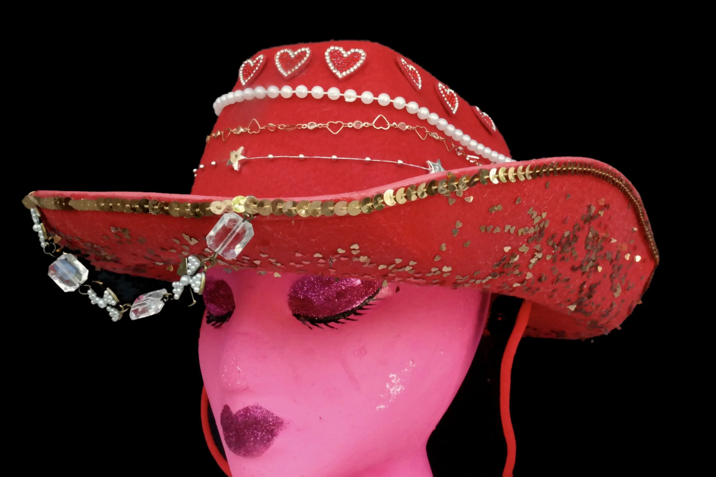 Pink mannequin head with glittery pink and purple eye makeup and lipstick, wearing a large red glittery wide-brimmed hat decorated with jewelry and hearts, against a black background.