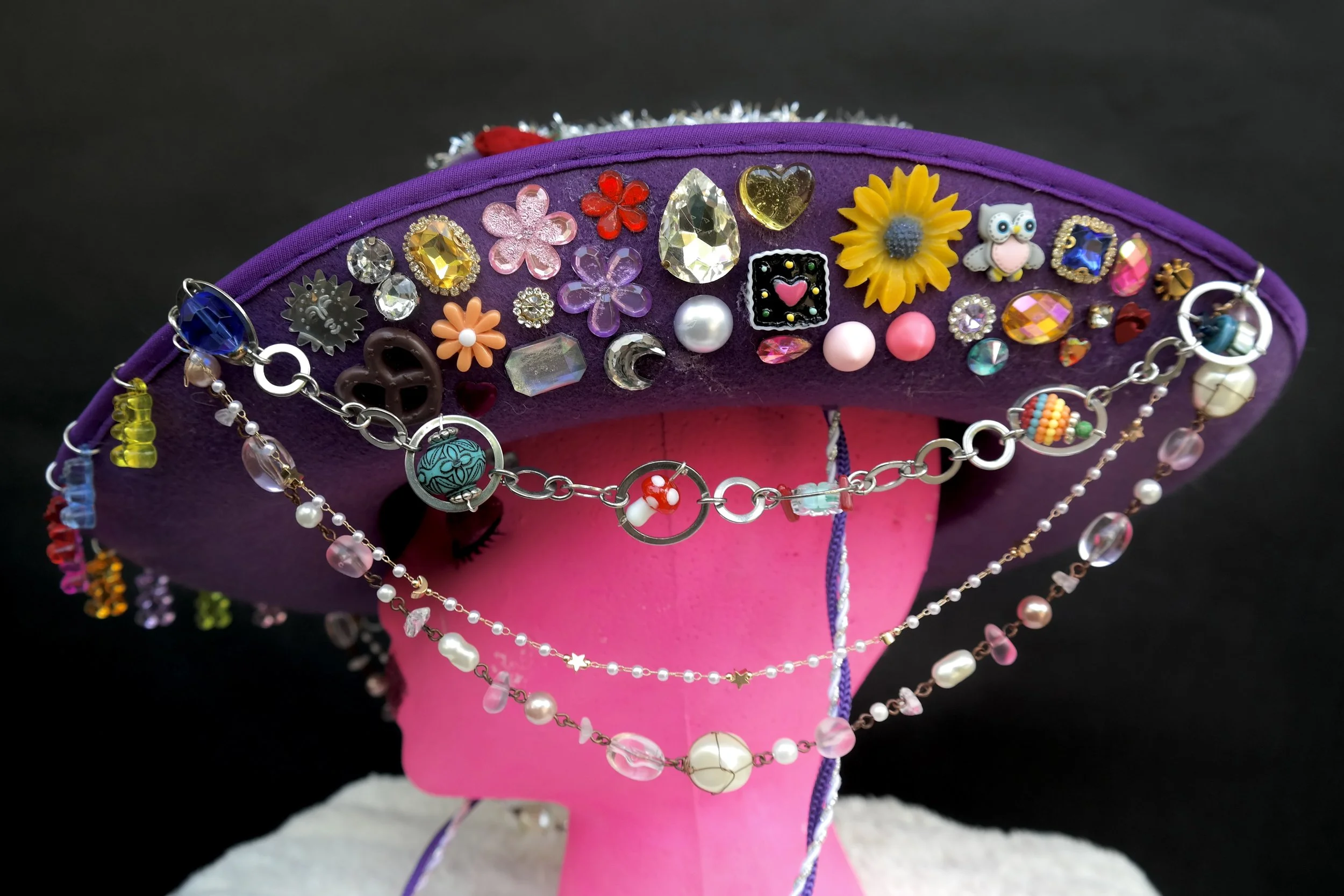 Purple hat decorated with various colorful pins, earrings, and jewelry pieces displayed on a pink mannequin head.