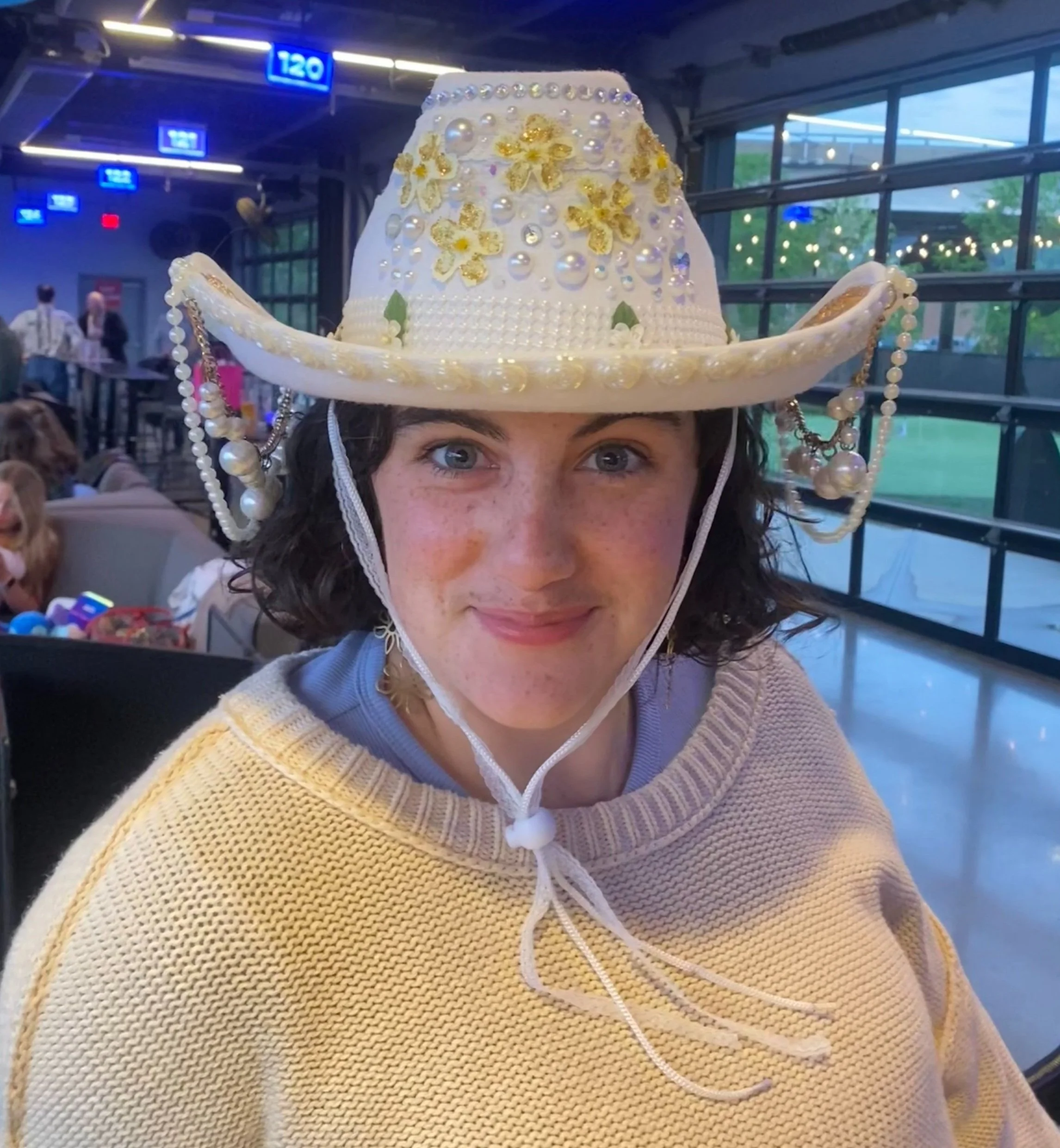 Woman wearing a decorated white cowboy hat with gold flowers, pearls, and beads, indoors at a social event.