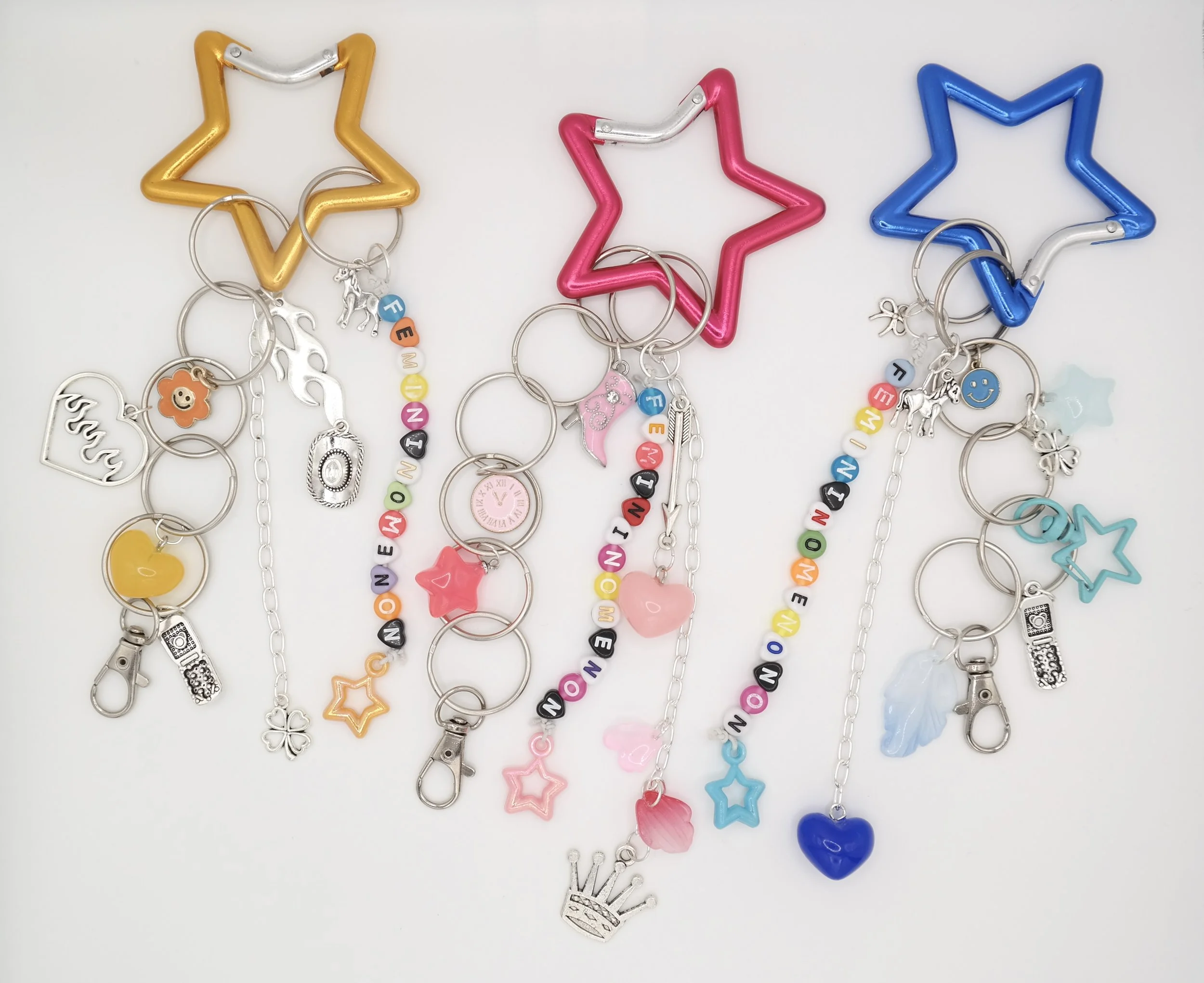 Four colorful star-shaped keychains with attached charm bracelets, each spelling out 'FEMINIST' with additional charms like hearts, stars, flowers, and smiley faces.