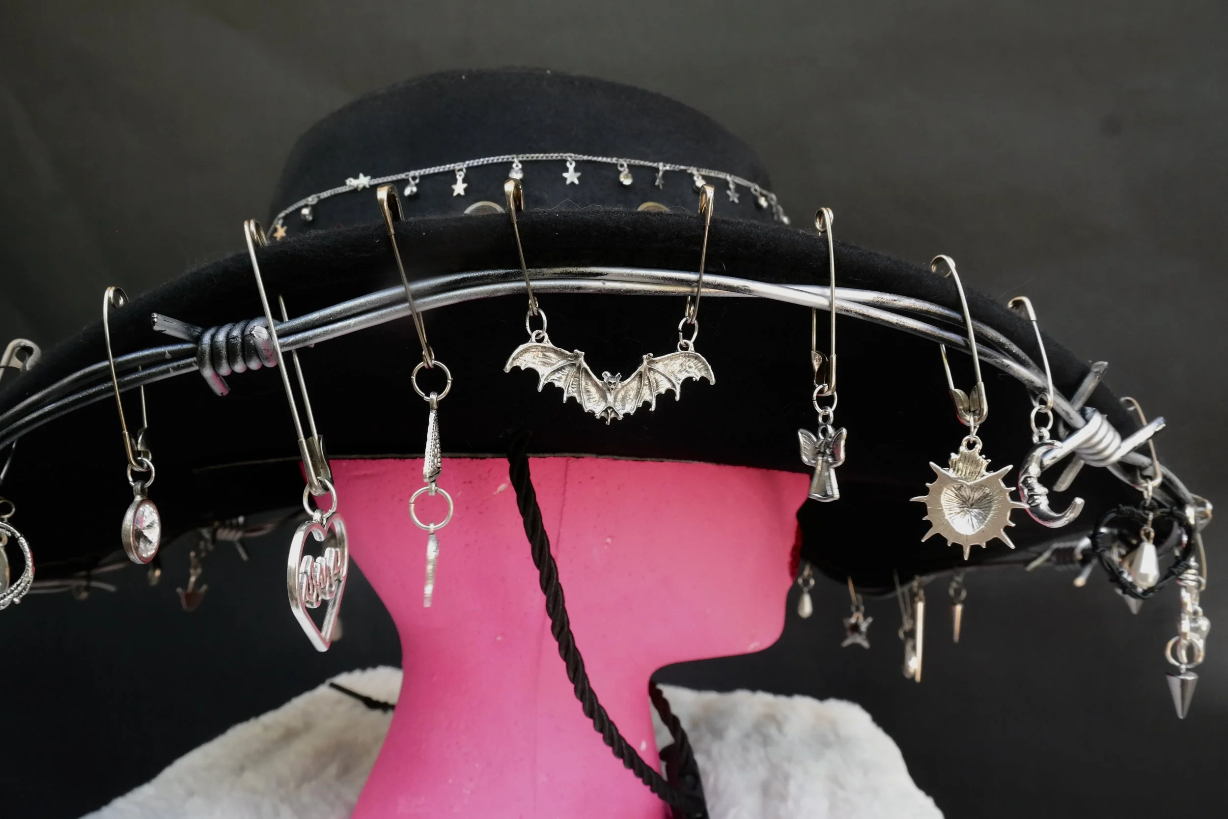 Black hat decorated with various silver charms and pins, displayed on a pink mannequin head with a white towel around its base.