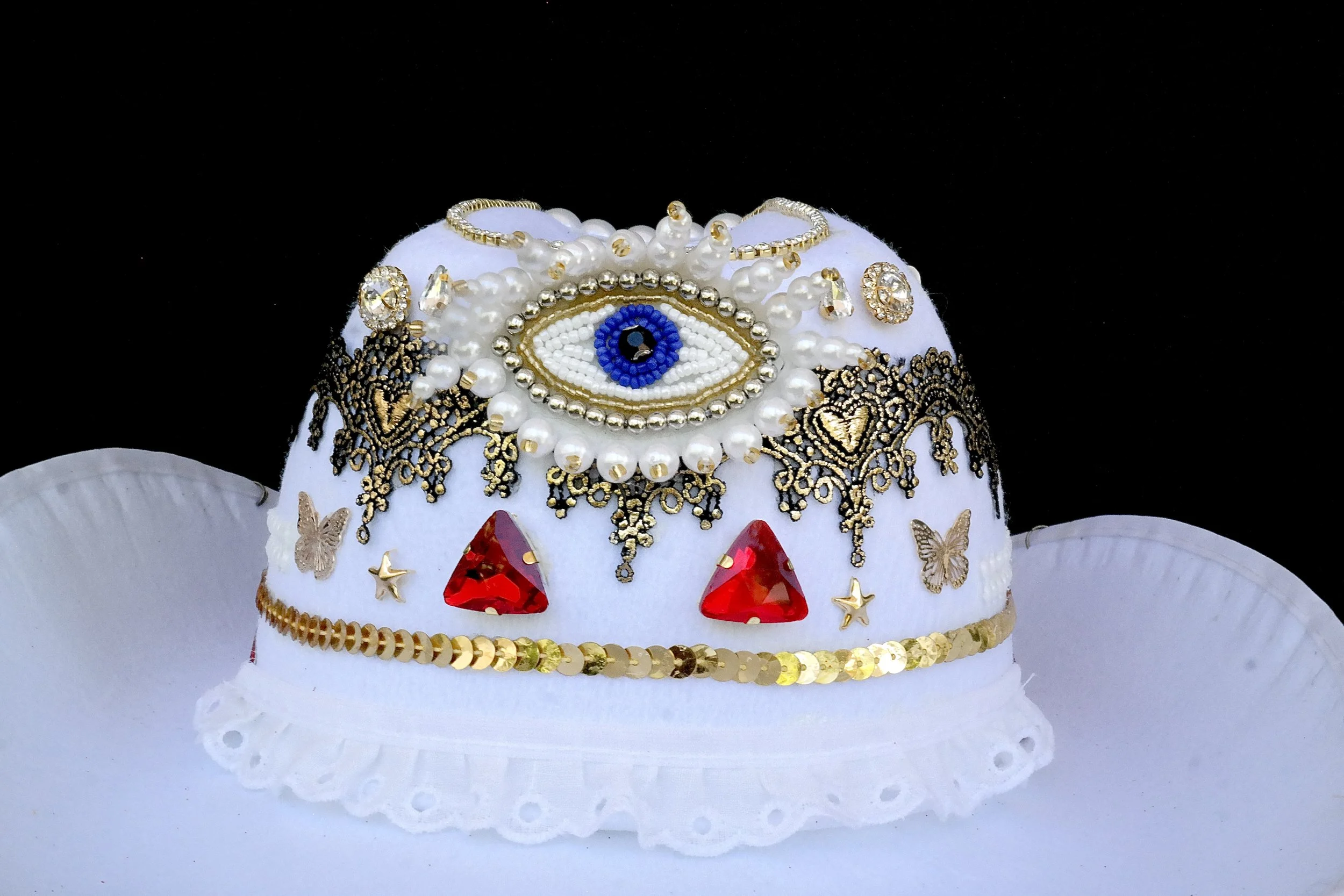 Decorative white hat adorned with jewelry, beads, and embroidery, featuring a prominent blue eye in the center, red gemstones, gold accents, butterflies, and small star-shaped embellishments, set against a black background.