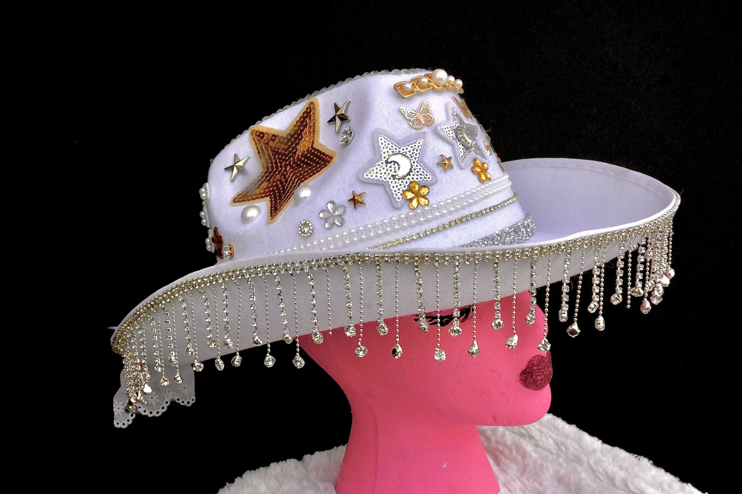 A white decorative hat with pearl and rhinestone embellishments, star and butterfly patches, and hanging rhinestone fringe on a pink mannequin head against a black background.