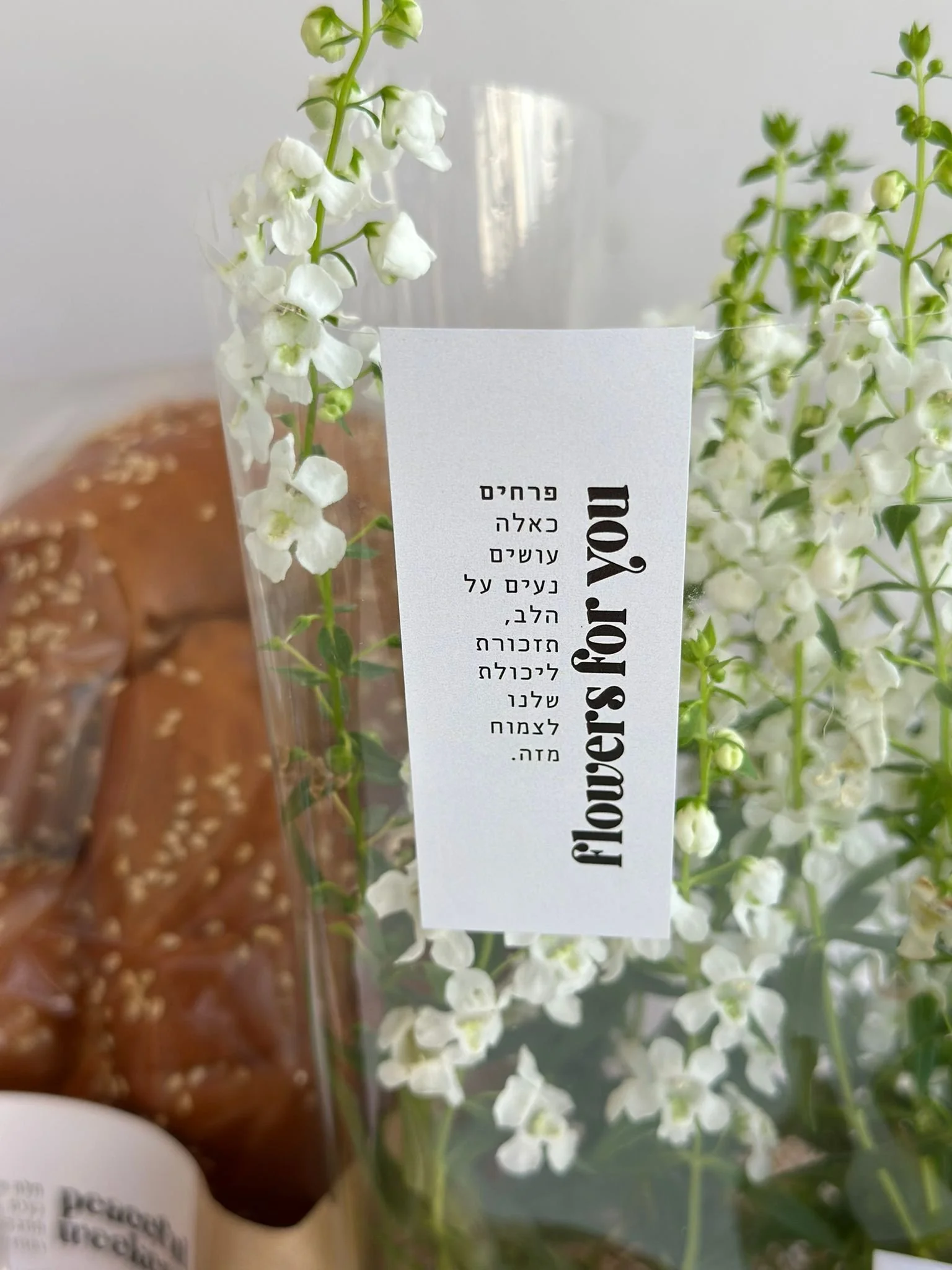 White floral bouquet with bilingual English and Hebrew tag reading “flowers for you,” styled beside a sesame-topped pastry — lifestyle image reflecting Jewish celebration and gifting.