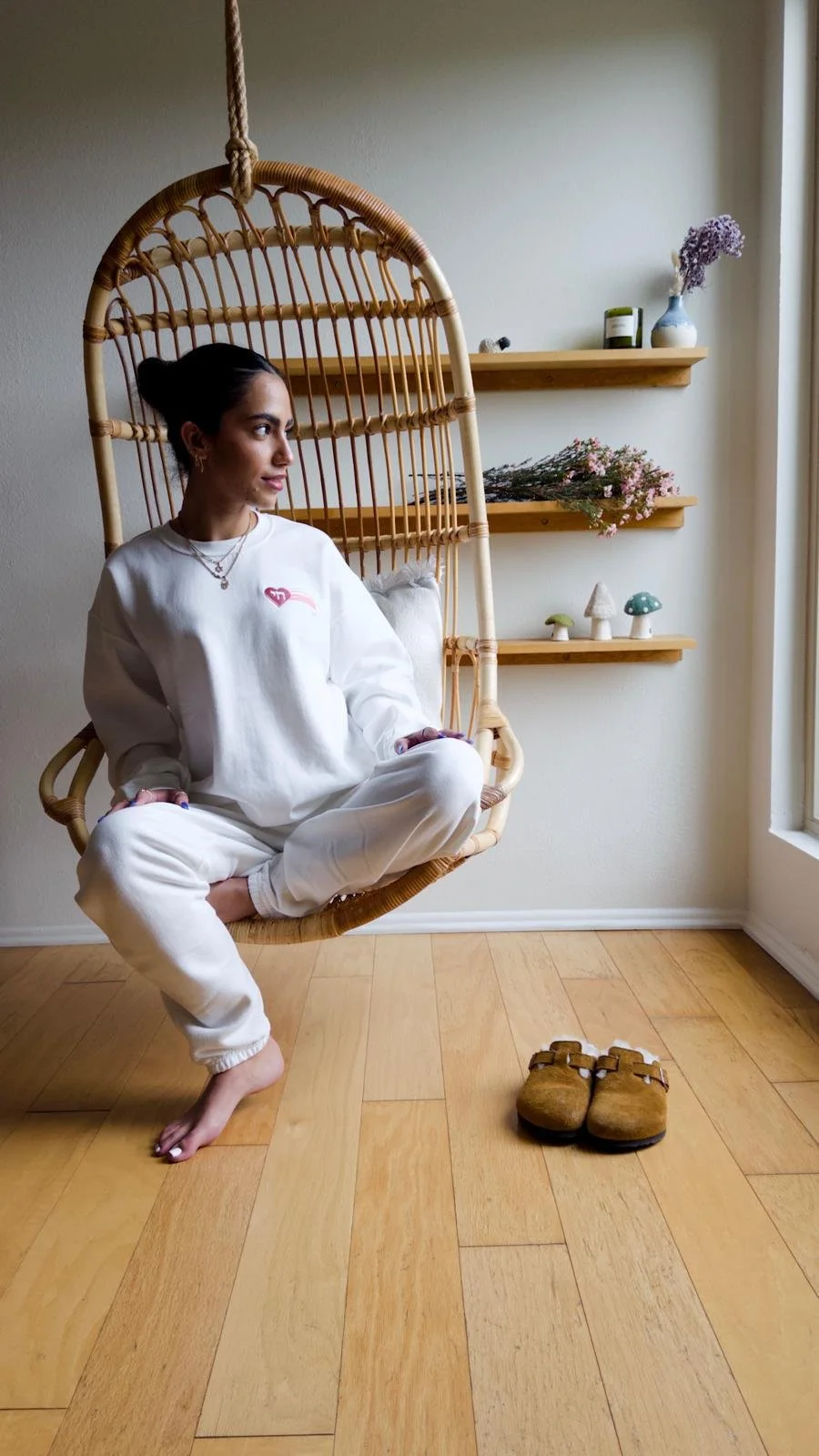 Woman relaxing in a hanging rattan chair near a window, styled in minimalist loungewear inside a light-filled room with natural wood floors.