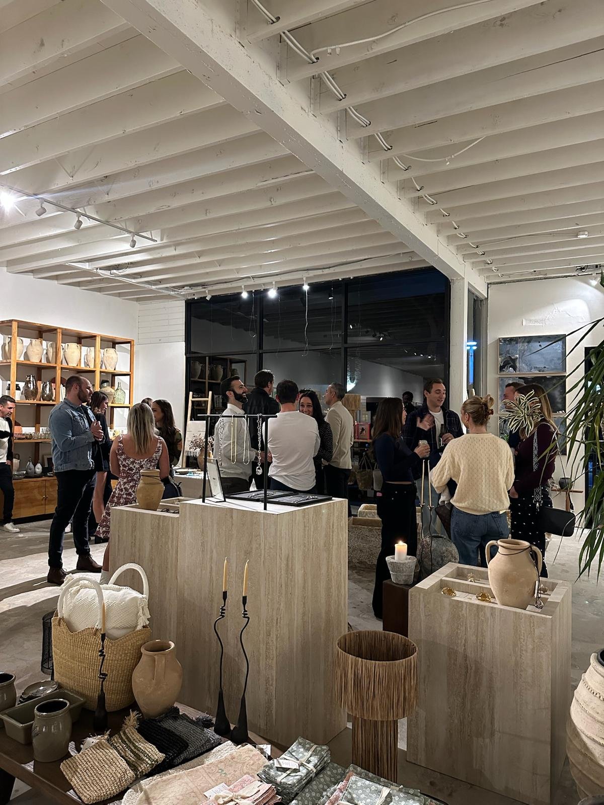 People gathered inside a modern retail store or gallery with pottery and home decor items on display. The store has a high, white painted wooden ceiling and large windows. Customers are browsing and conversing.