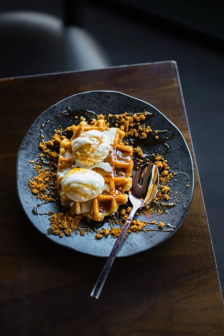 Waffle topped with ice cream, caramel sauce, and crumbled toppings on a black plate, with a spoon on a wooden table.