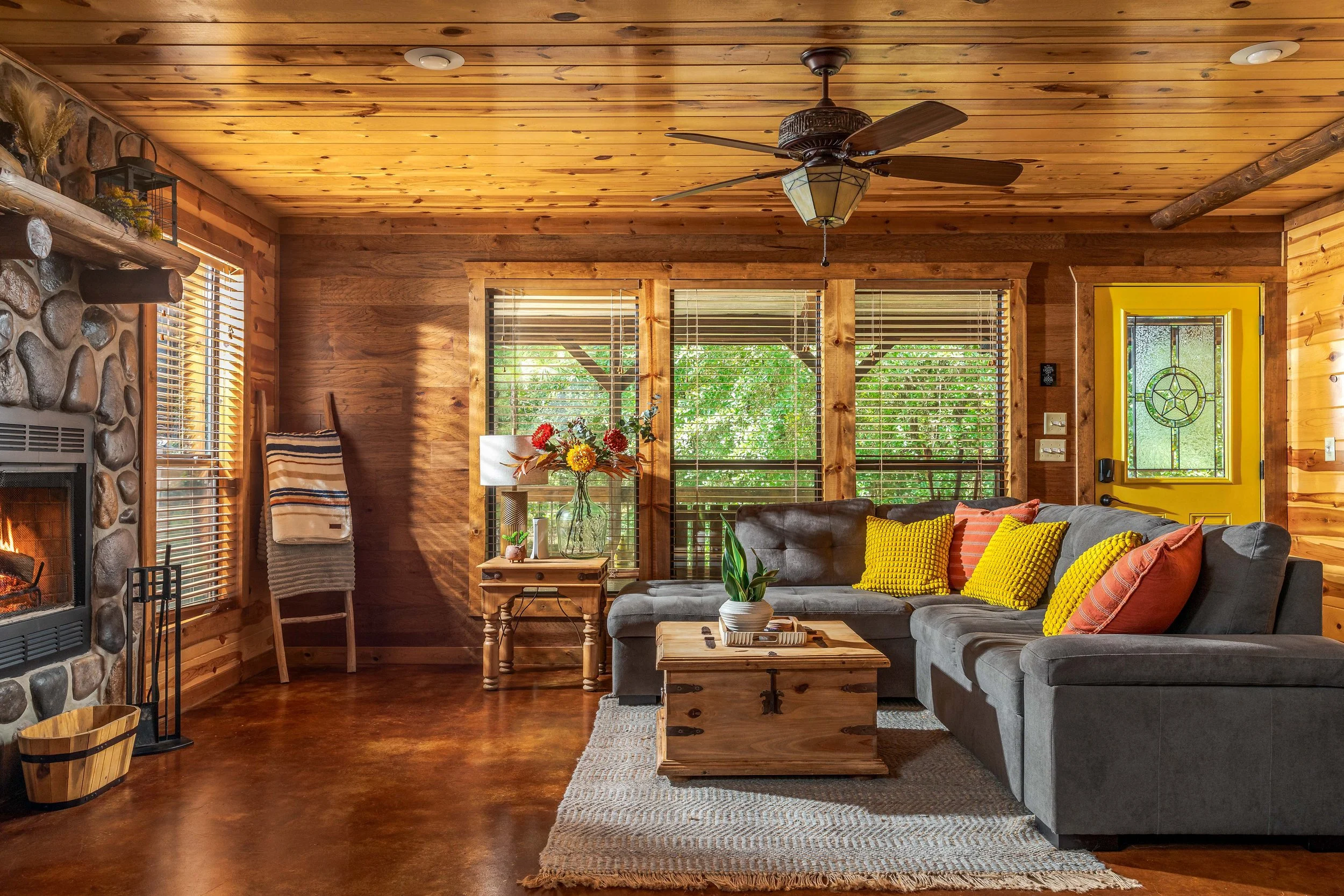 Living room with wood-paneled walls, stone fireplace, gray sectional sofa with yellow and orange pillows, wooden coffee table with a plant, large windows with blinds, and a yellow front door with glass and decorative star design.