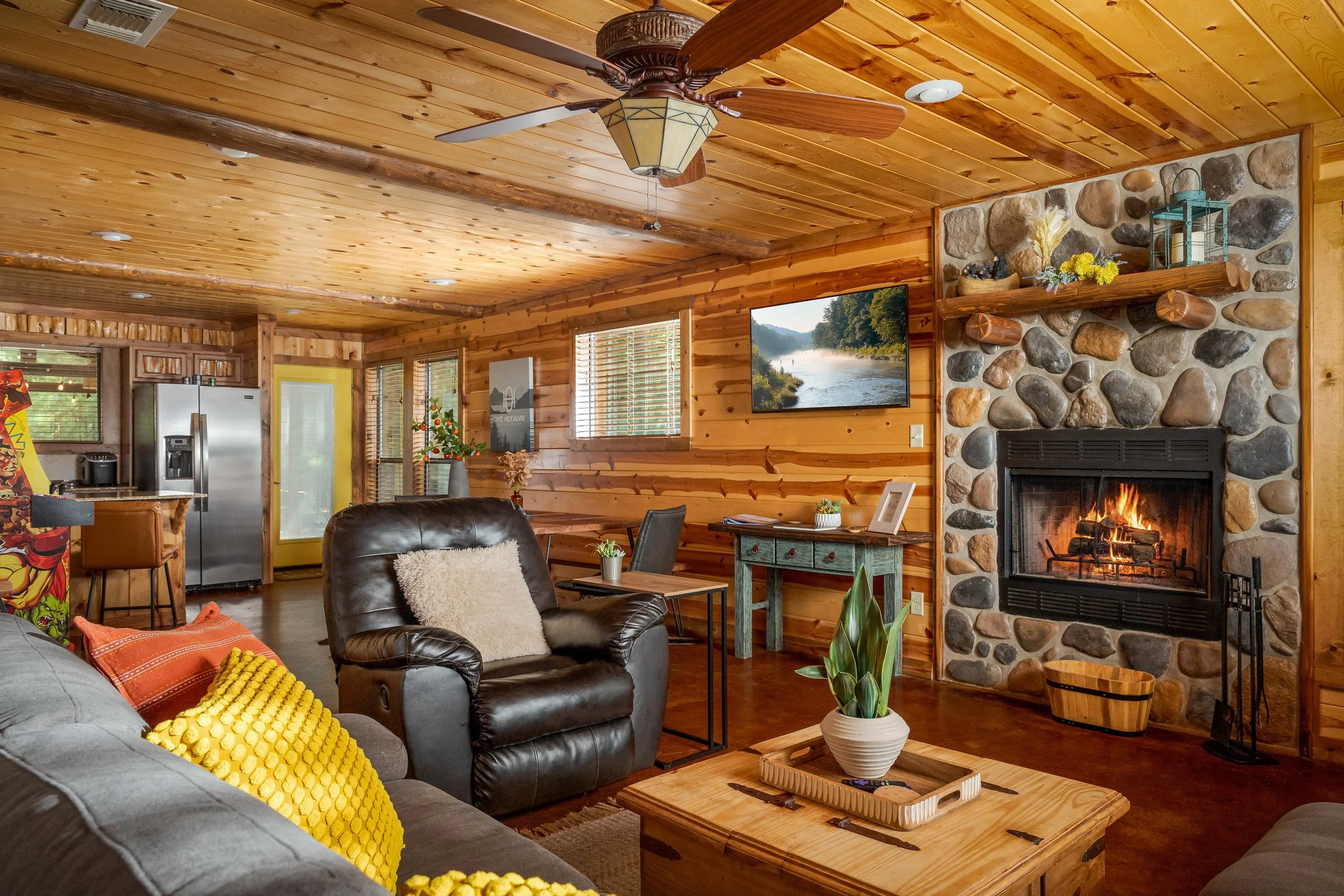 Cozy living room with wooden walls and ceiling, stone fireplace with shelf, leather and fabric couches, coffee table with plant, wall-mounted TV, and windows with blinds in a cabin-style home.