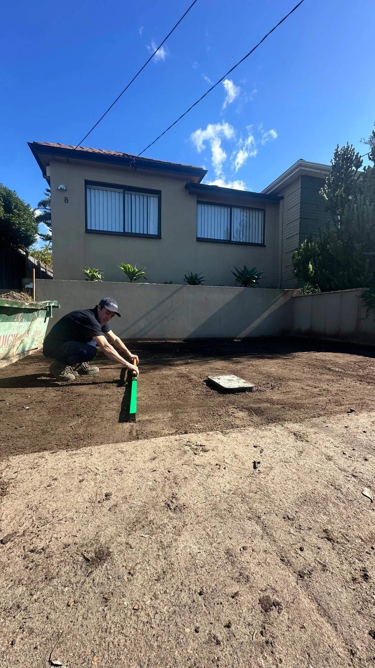 A man in a black shirt, black pants, and a black cap squats on a dirt lot while using a tool to level or measure the ground. There is a house with large windows and green plants in the background, under a blue sky with some clouds.