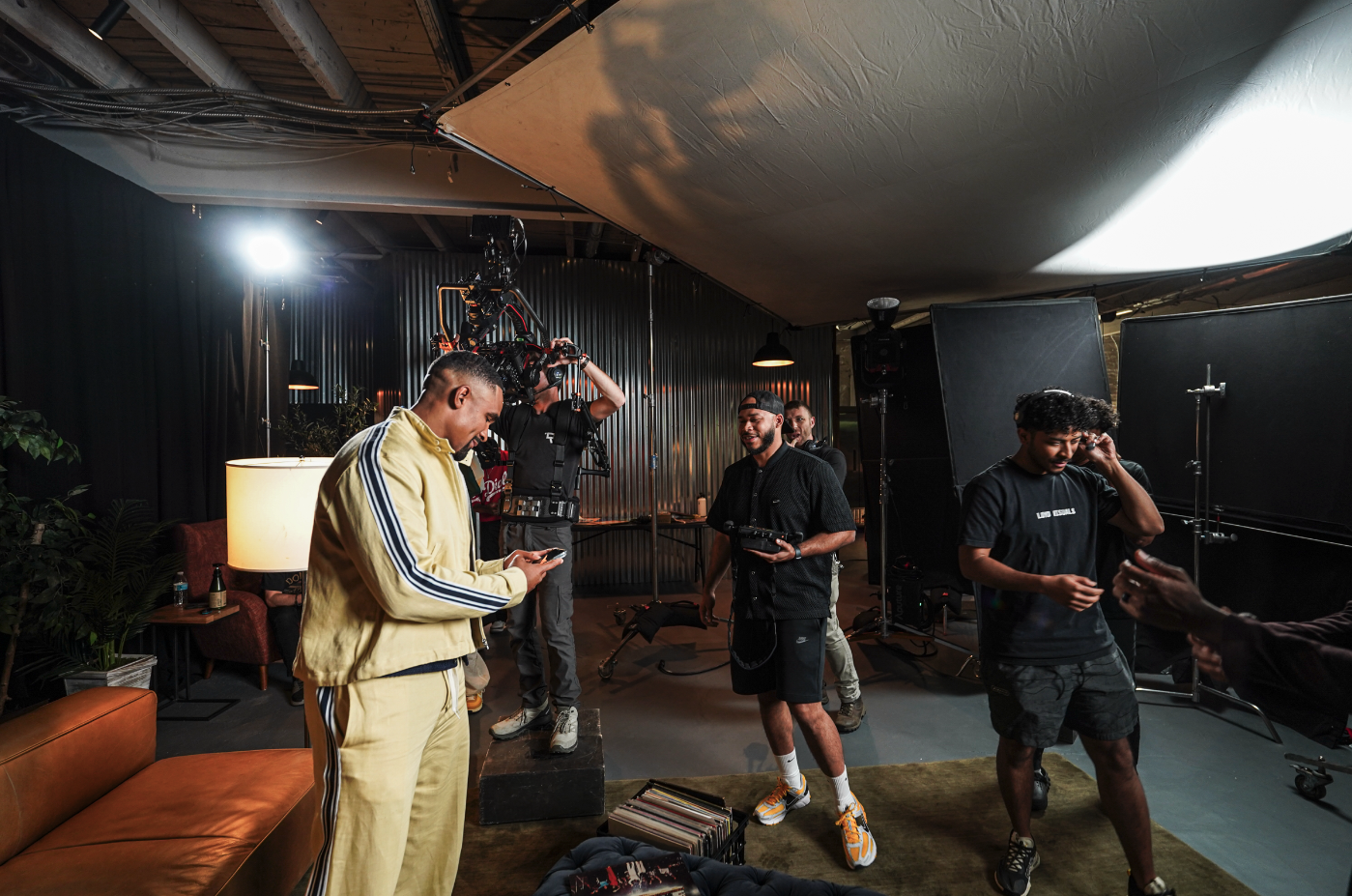 A behind-the-scenes look at a film or video production set, featuring a group of people. One man in a yellow tracksuit checks his phone, while others handle camera equipment and lighting. The setting includes various lights, a large rig, and a sofa in a casual studio environment.