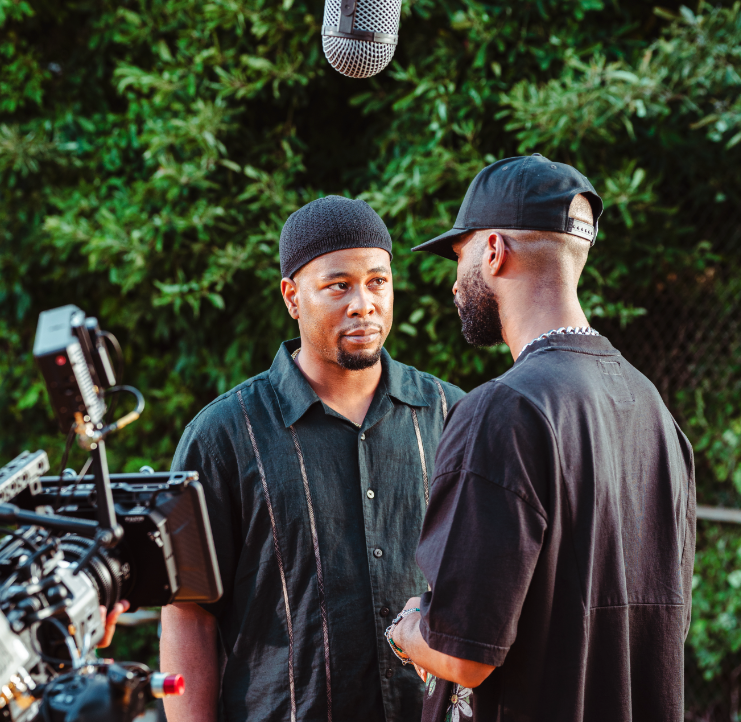 Two men interacting on a film set, with a camera and boom microphone visible, outdoors with greenery in the background.