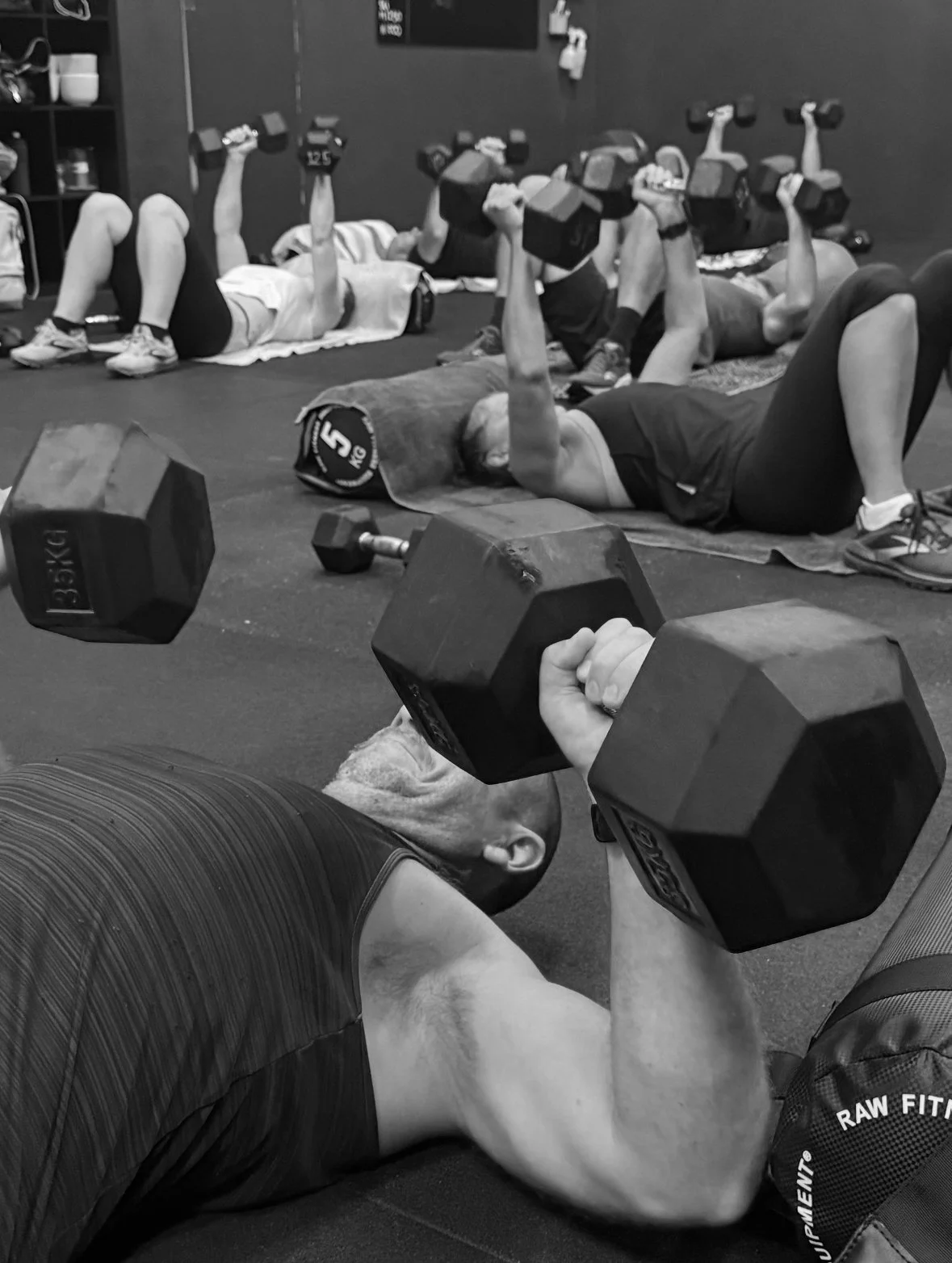 People lying on exercise mats lifting dumbbells in a group fitness class at a gym. A close-up shows a man in the foreground holding dumbbells.