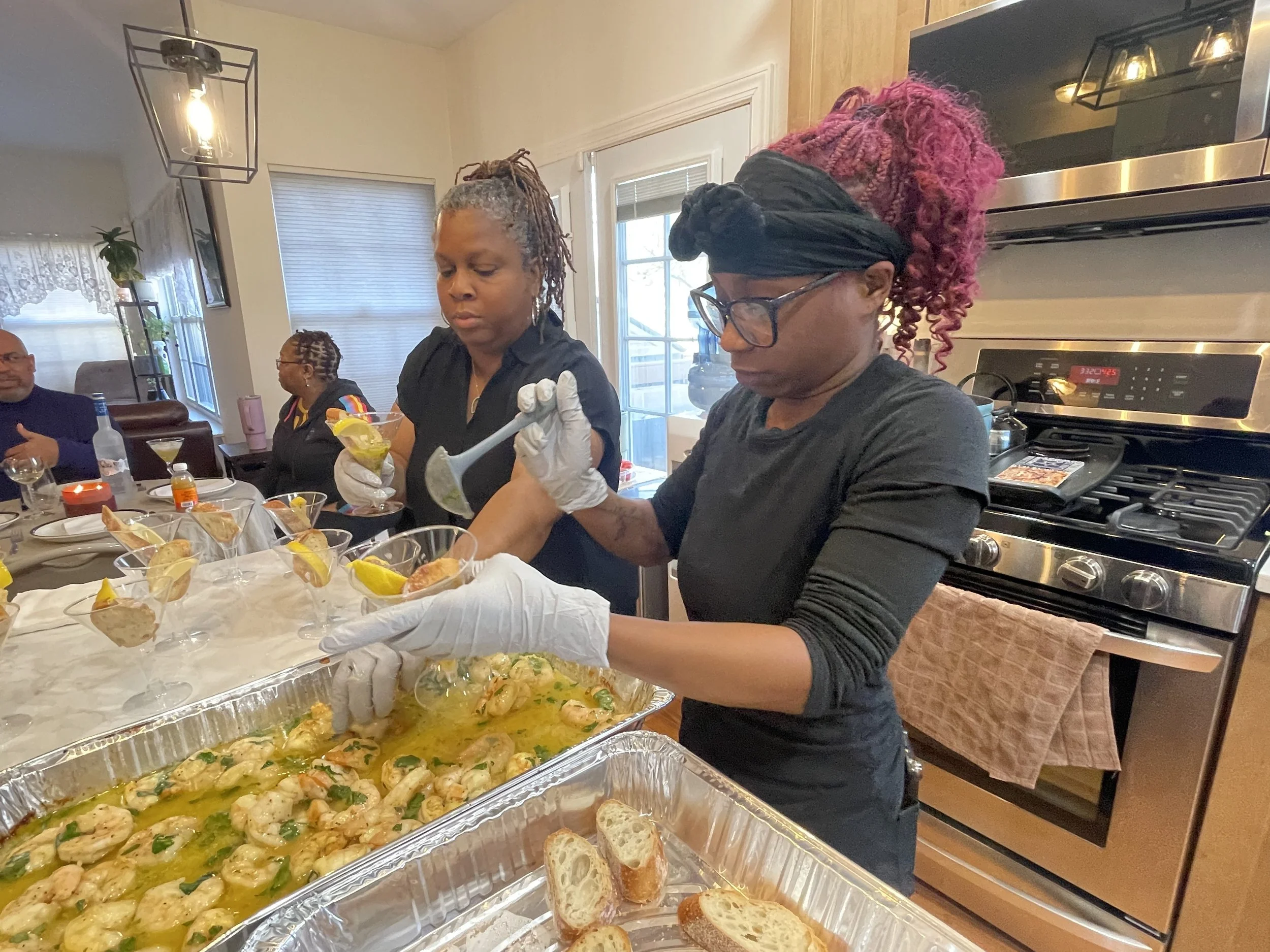 Two women preparing shrimp dishes at a kitchen counter, with guests seated at a dining table in the background.
