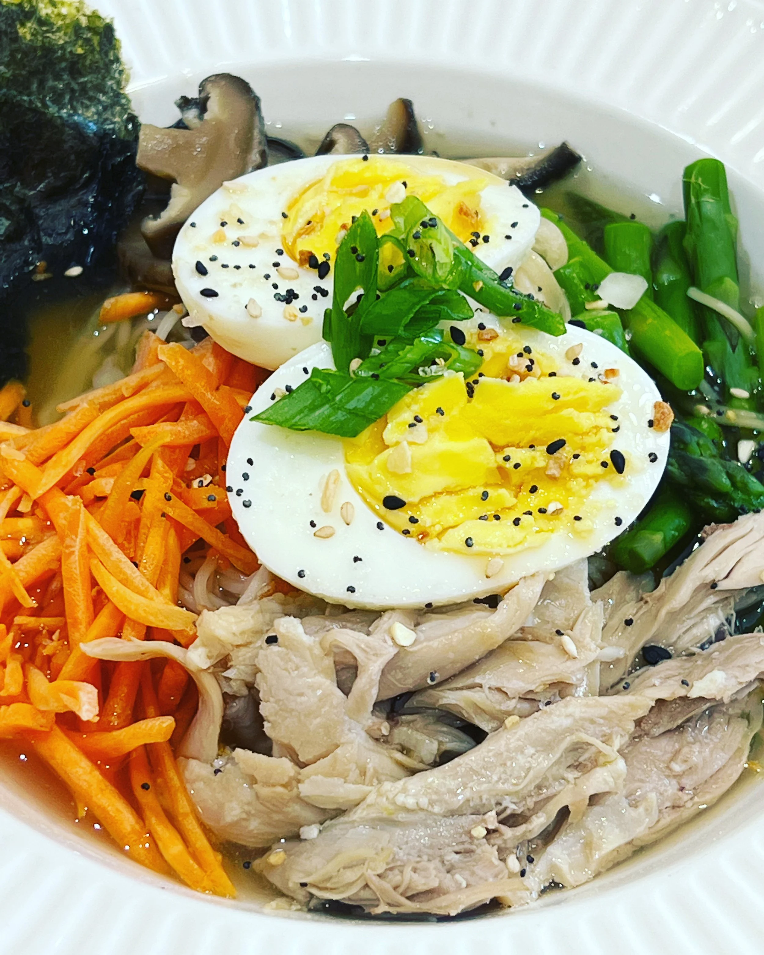 A bowl of Korean-style noodle soup with shredded chicken, shredded carrots, chopped green onions, halved boiled eggs, seaweed, and bean sprouts, garnished with sesame seeds and black sesame seeds.